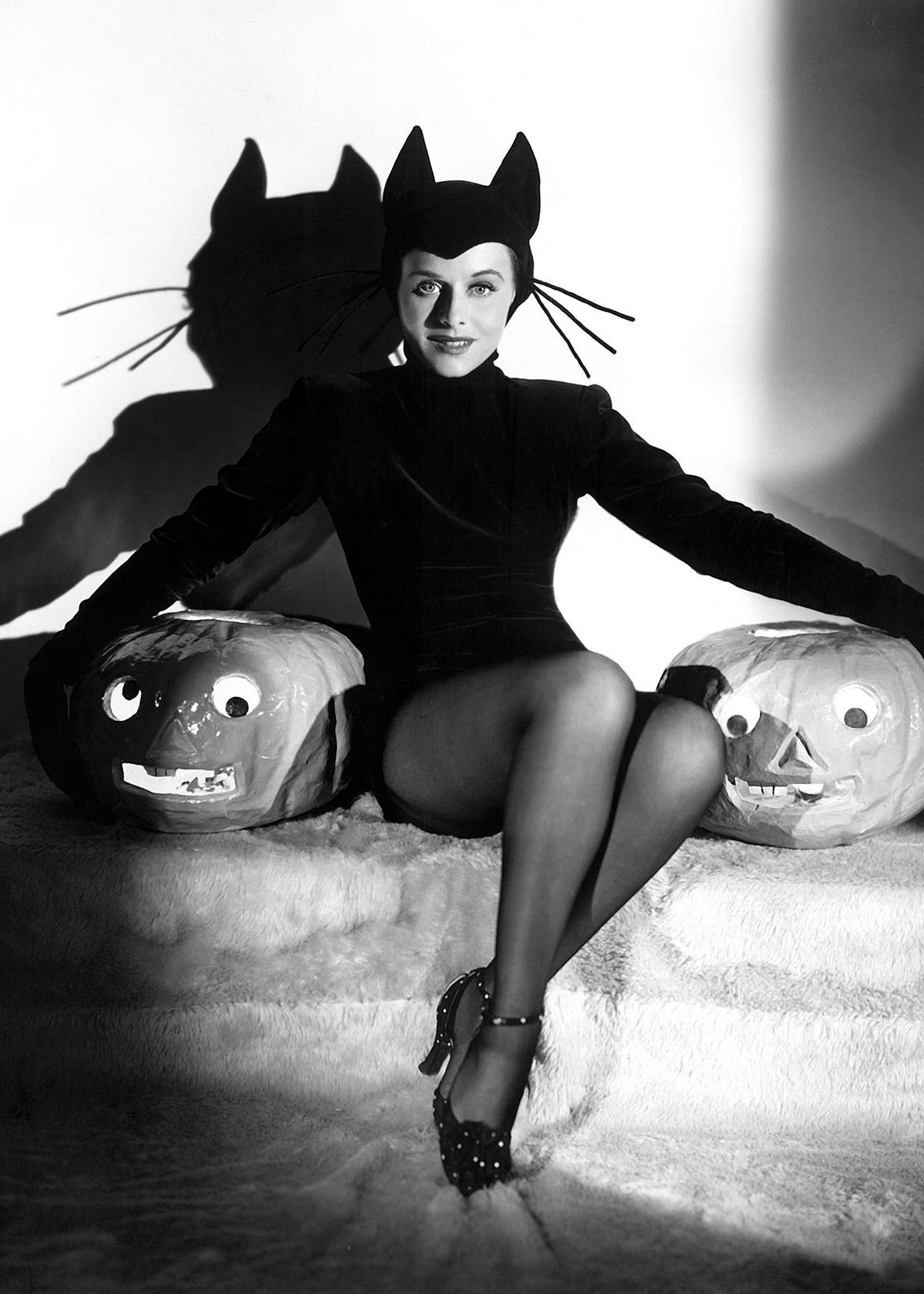 An actress is in a cat costume, flanked by two prop jack-o’-lanterns