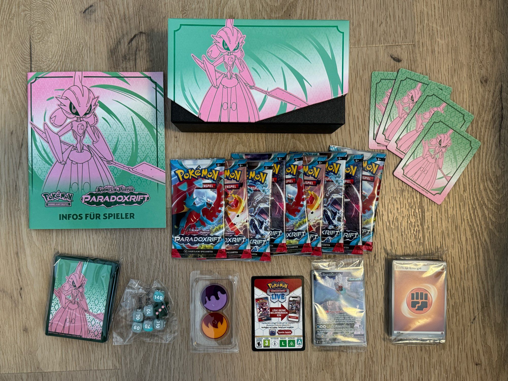 Inhalt der Top-Trainer-Box „Karmesin & Purpur – Paradoxrift: Eisenkrieger“: Sammelbox, neun Booster, Kartenhüllen, Würfel, Marker, Energiekarten, Trennstege, Promokarte, Codekarte und Regelhelft.