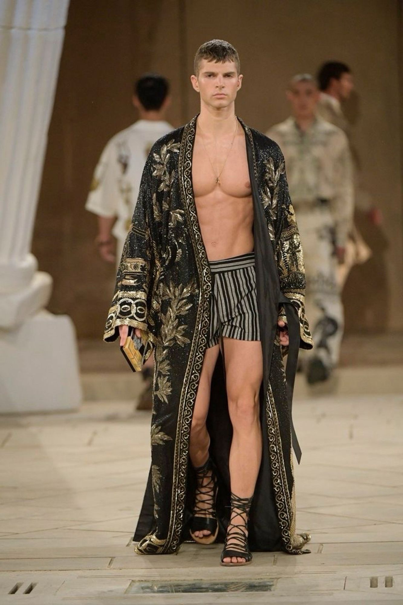 A male model from the Dolce&Gabbana Alta Sartoria Menswear Show. He is wearing a black robe embroidered with gold, black-and-grey vertically striped shorts, and black gladiator-style sandals.