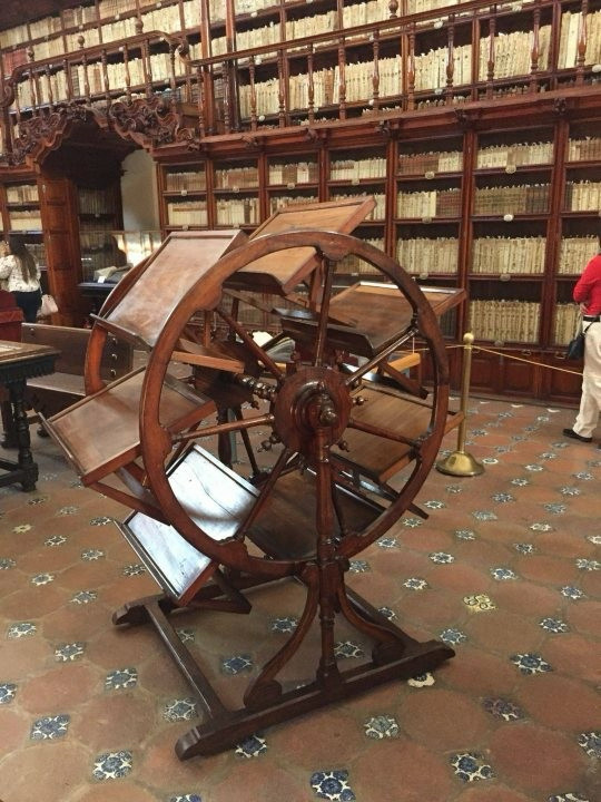 A photo of an antique wooden machine. It has a series of wooden slats between two wheels which  can be rotated to keep te slats in a readable position. It’s situated in a an ancient old wooden library.