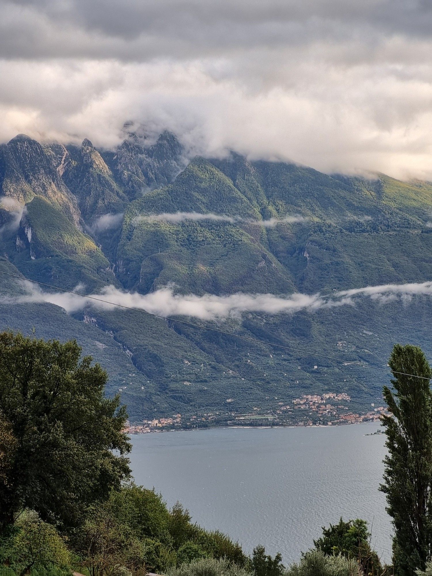 Blick auf den Gardasee, eingerahmt von Bäumen, gegenüber eine Bergkette mit Wolkenband