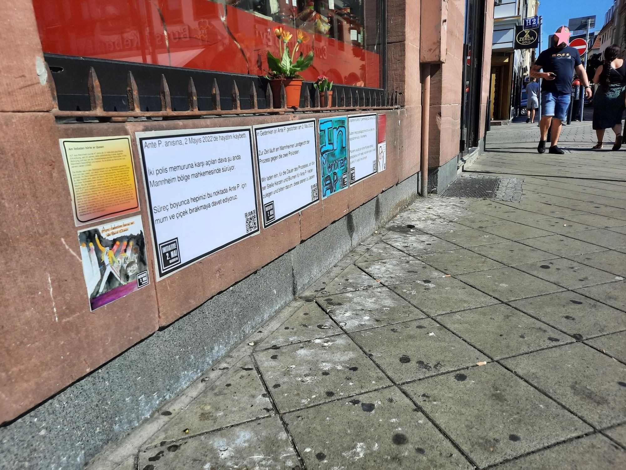 Auf dem Bild: Der Bürgersteig, darüber die Mauer unterhalb des Schaufensters der Metzgerei am Marktplatz. An der Mauer sind Plakate in verschiedenen Sprachen angebracht, die Informationen zu Ante P. enthalten. Am oberen, äußeren Rand der Mauer, vor dem Schaufenster befindet sich ein Metallsteg, an den Zacken nach oben gehen. Dahinter das Schaufenster welches am unteren Rand von innen mit roter Folie abgedeckt ist. Darüber erkennbar Lebensmittel. Davor, also zwischen Metallbalken und Schaufenster stehen 2 x Blumen im Topf. Auf dem Bürgersteig Menschen, die auf die Metzgerei zugehen.