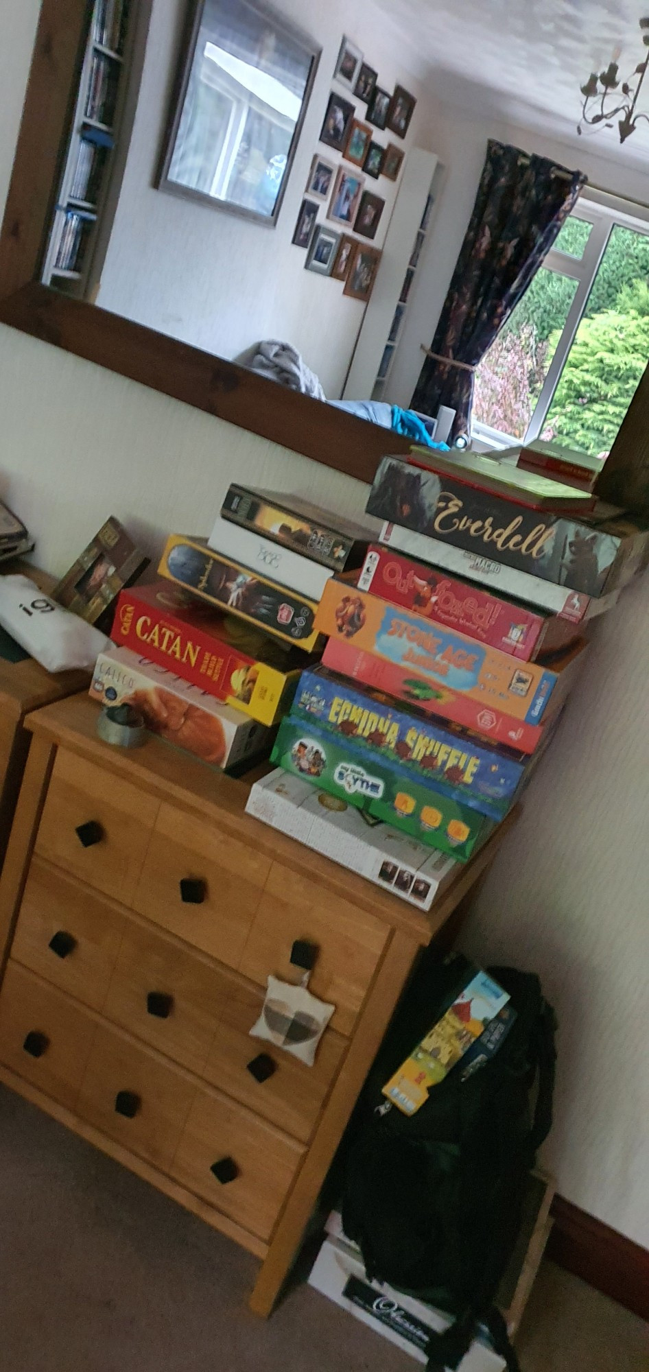 A set of living room drawers with a pile of games on it. A bag sat next to it with a boardgame sticking out of it. Under the bag are two boardgames laying on the floor.... 