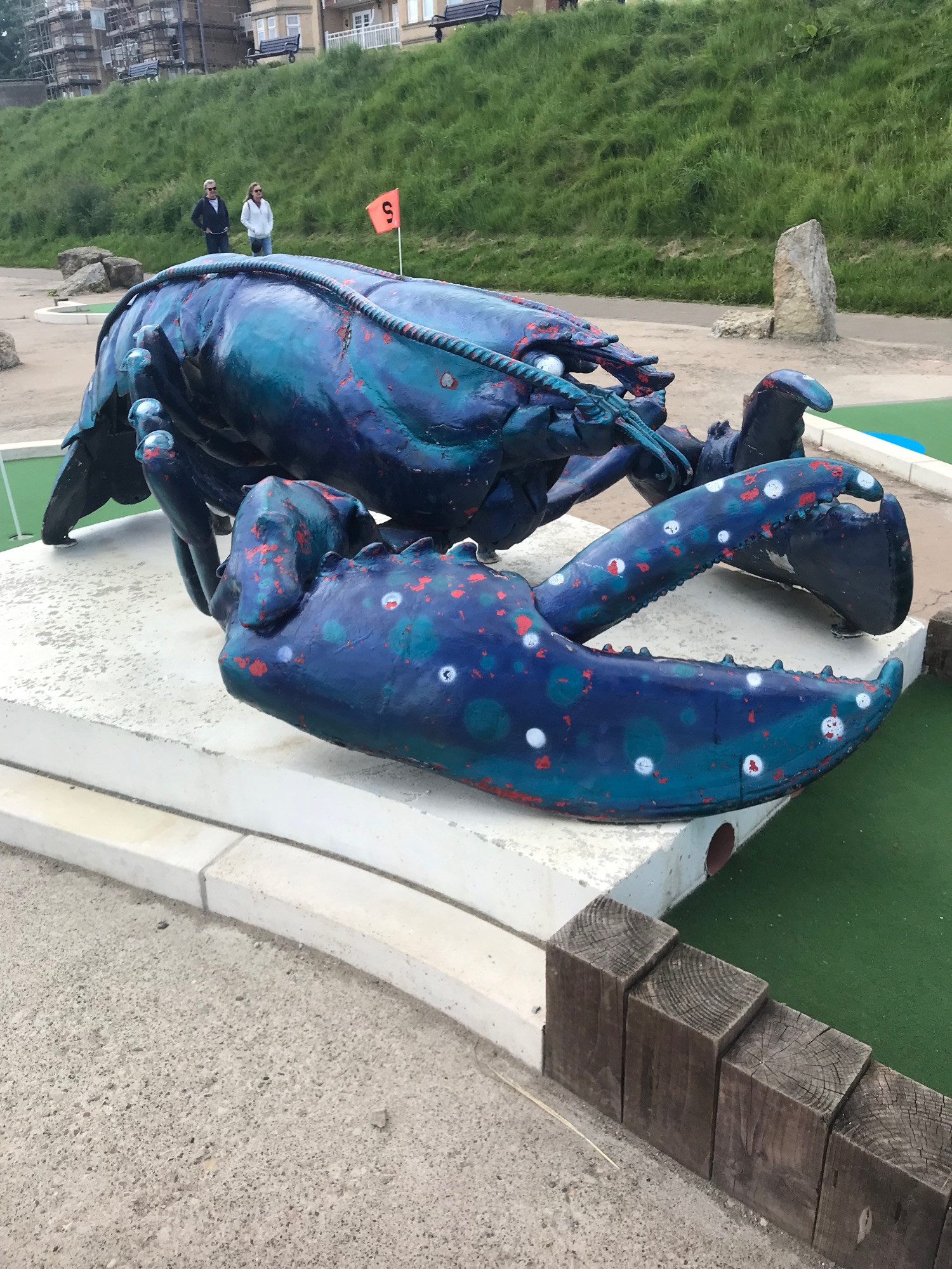 A large model of a blue lobster with stickers on its claws on a white plinth, it is part of a crazy golf course .

