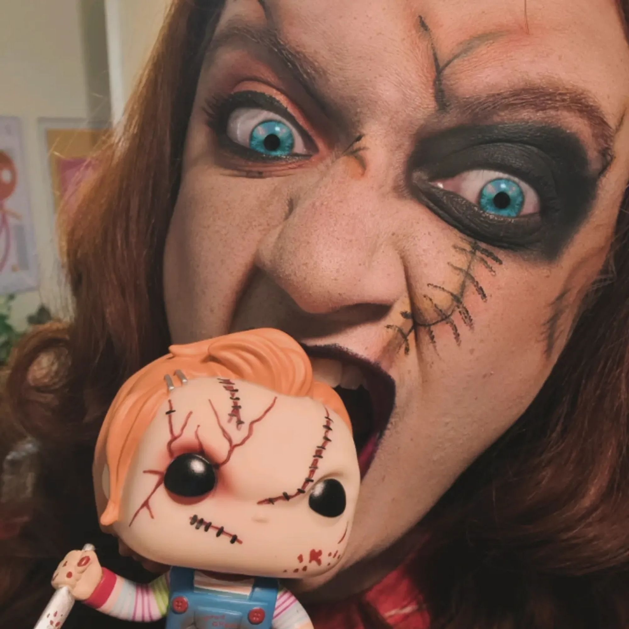 Eu, com uma maquiagem no rosto que imita os cortes e remendos do personagem Chucky, da série de filmes O Brinquedo Assassino. Seguro um bonequinho funko pop do Chucky e mordo sua cabeça.