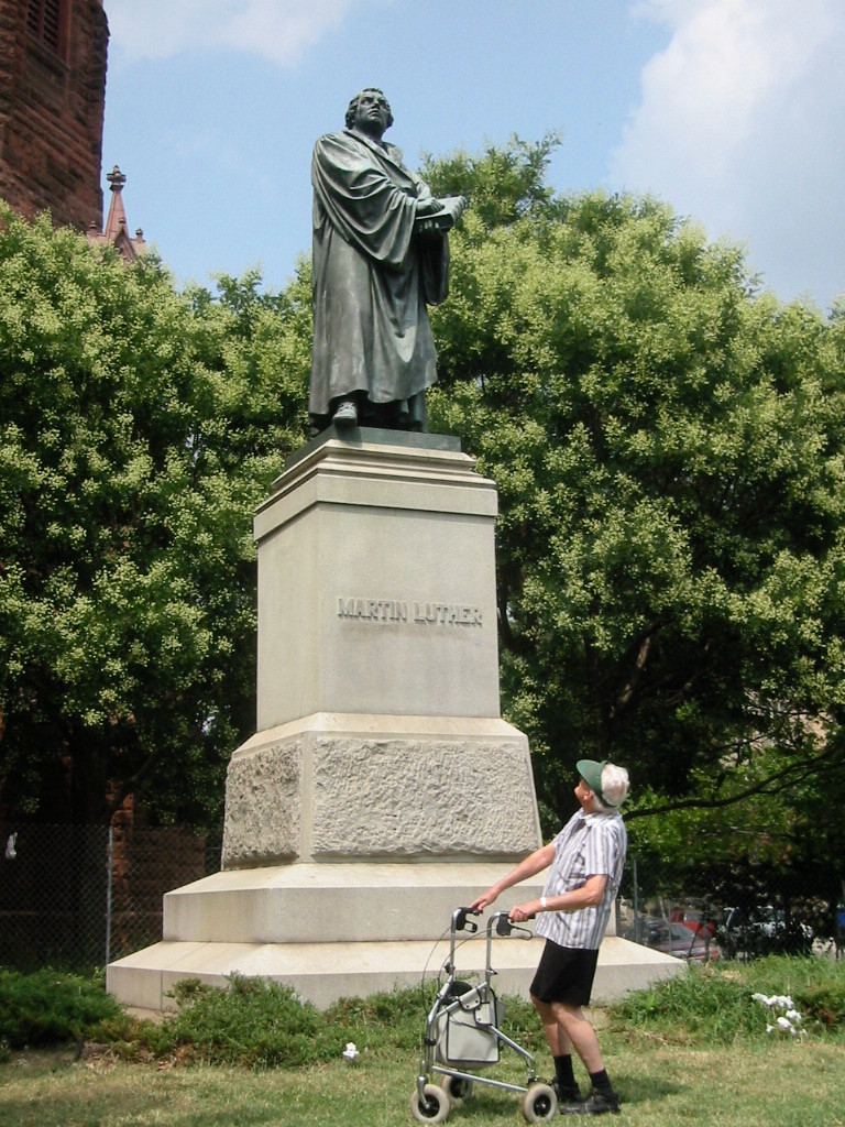 An elderly man with a walker, wearing shorts, a short-sleeved shirt, and a cap, stands on a lawn below the statue of Martin Luther in front of Luther Place Memorial Church at Thomas Circle in Washington, DC. The statue is on a high pedestal and the man is leaning back and looking up at it. Behind the statue is a tree with dense foliage. A portion of the dark red church building is visible at the far left.