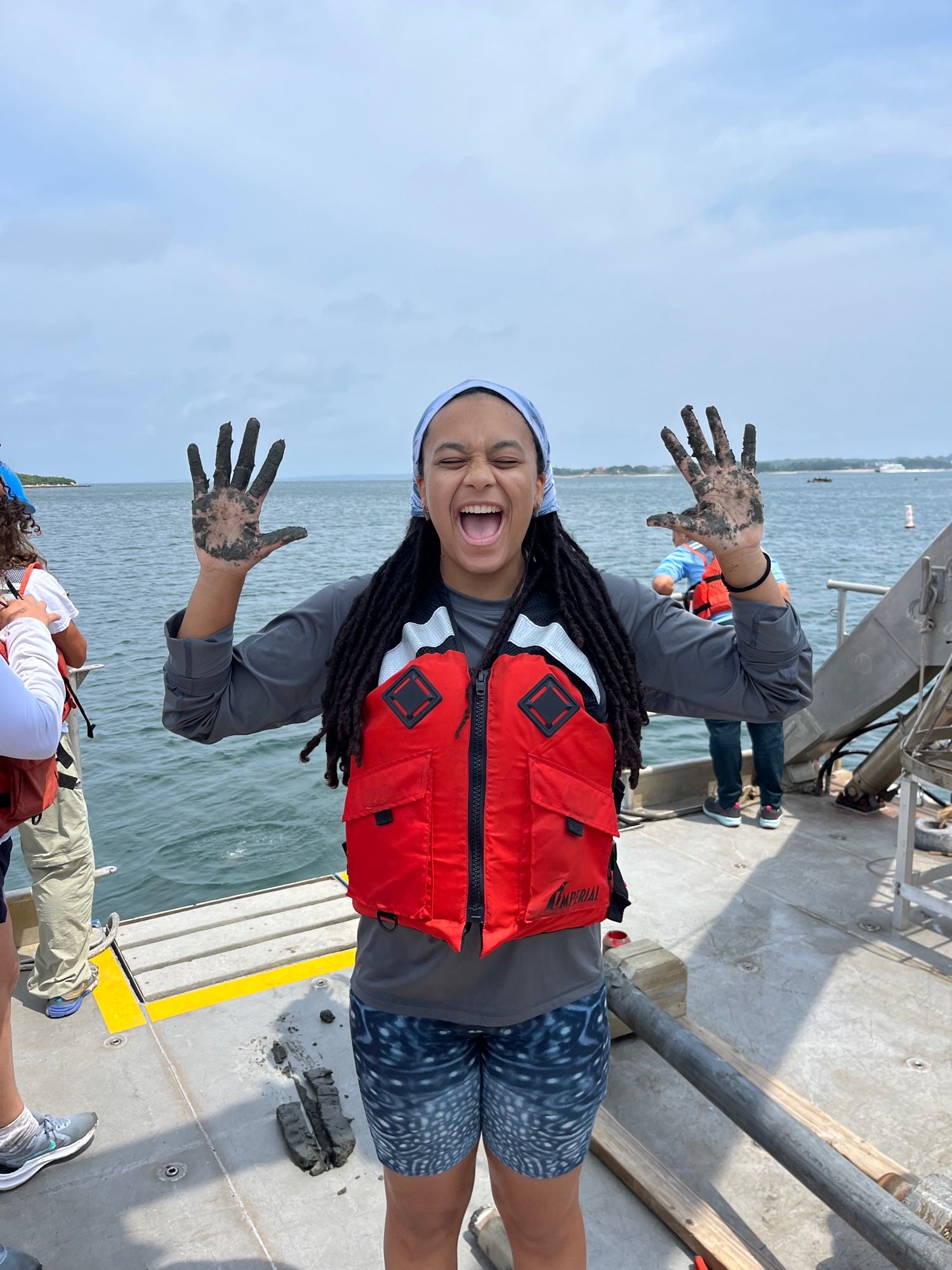 Jaida in a boat doing oceanography activities with sediment in her hands. She is wearing a bright orange life vest and whale shark shorts!