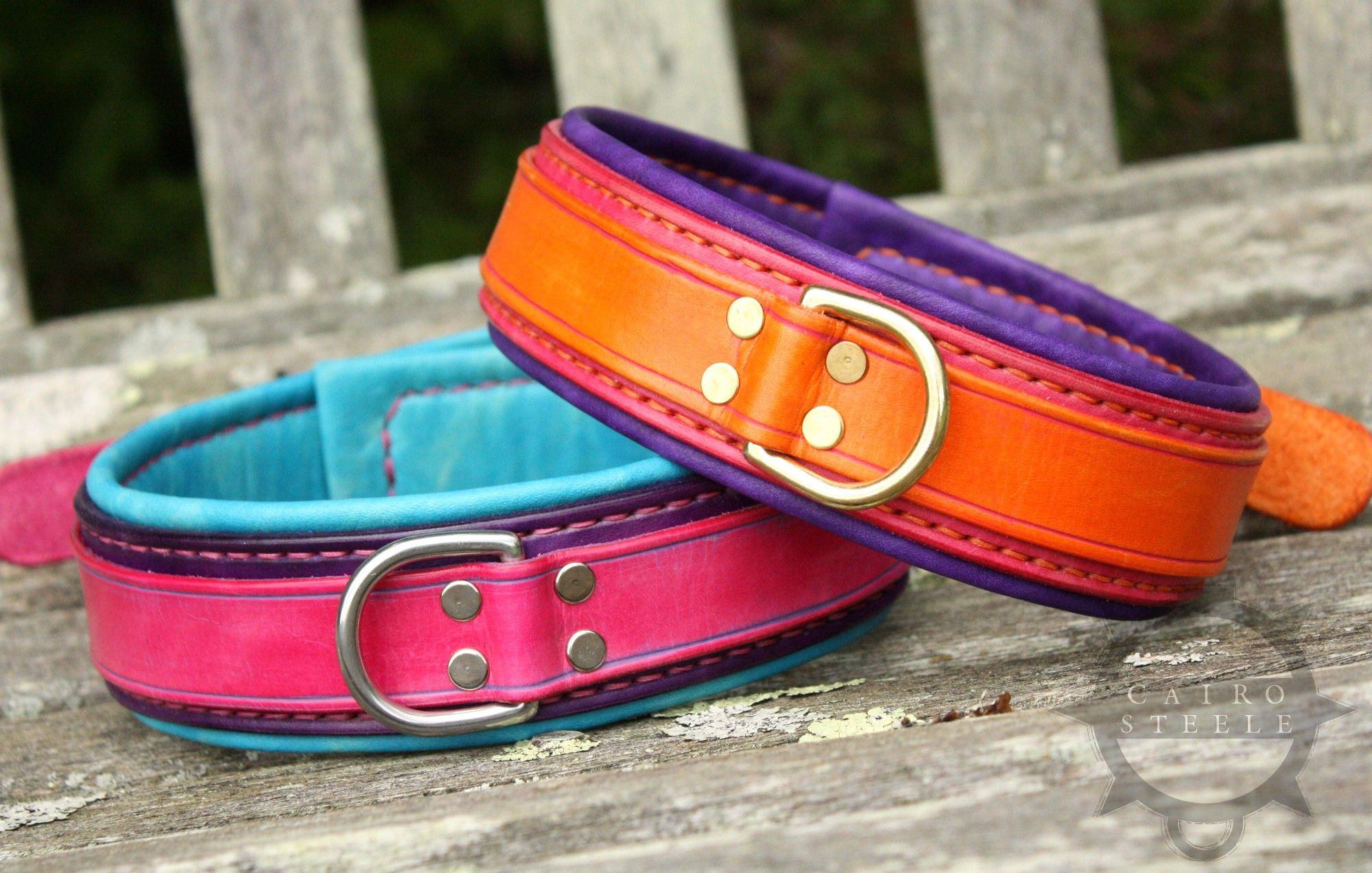 A pair of collars photographed at an angle. The top collar is orange, pink, and purple, with orange thread and brass hardware. The bottom collar is pink, purple, and blue, with pink thread and stainless steel hardware.