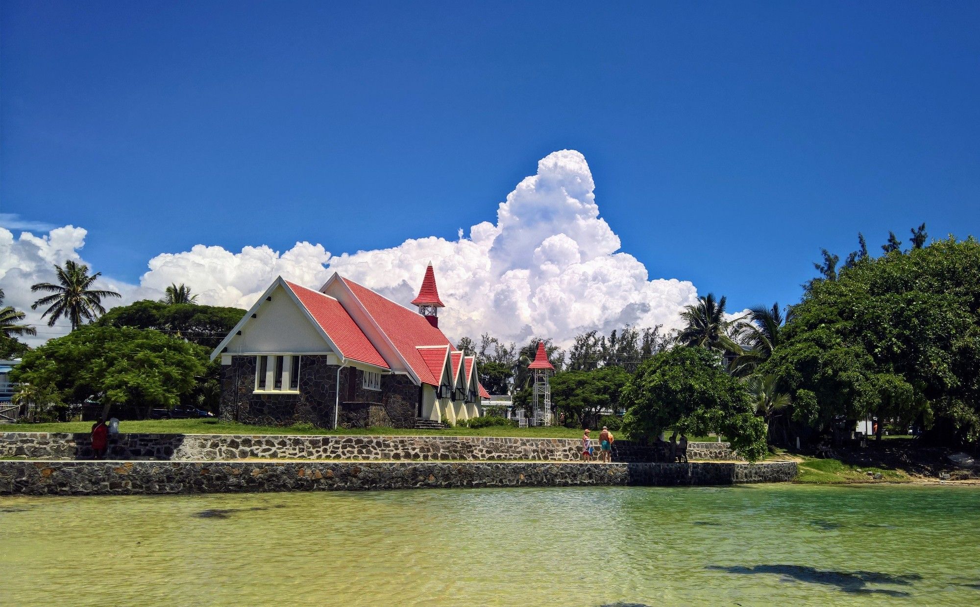 Eine Holzkirche mit rotem Dach steht direkt am Strand. Das Meer ist türkis, links und rechts der Kirche stehen Bäume. Ich kann Palmen erkennen. Im Hintergrund der blaue Himmel aus dem eine riesige weiße Gewitterwolke emporsteigen.