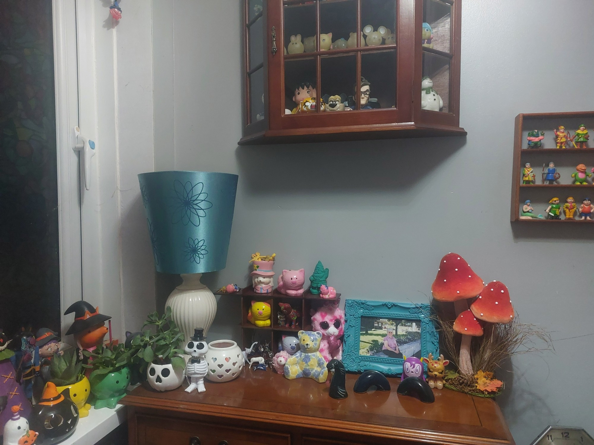 The cabinet at the top of my stairs, complete with artificial toadstools, a framed photo of my mum, 3/4 of a vintage Loch Ness monster figure, a ceramic teddy bear, a skeleton bobblehead, a tealight holder full of rubber balls, a plant growing out of a skull and a lamp with an upside down shade. Above it is a large-ish wall until filled with various toys and figures and a small set of shelves filled with vintage Soma figures. To the left is a windowsill covered in Halloween decorations and more plants, this time in octopus pots.