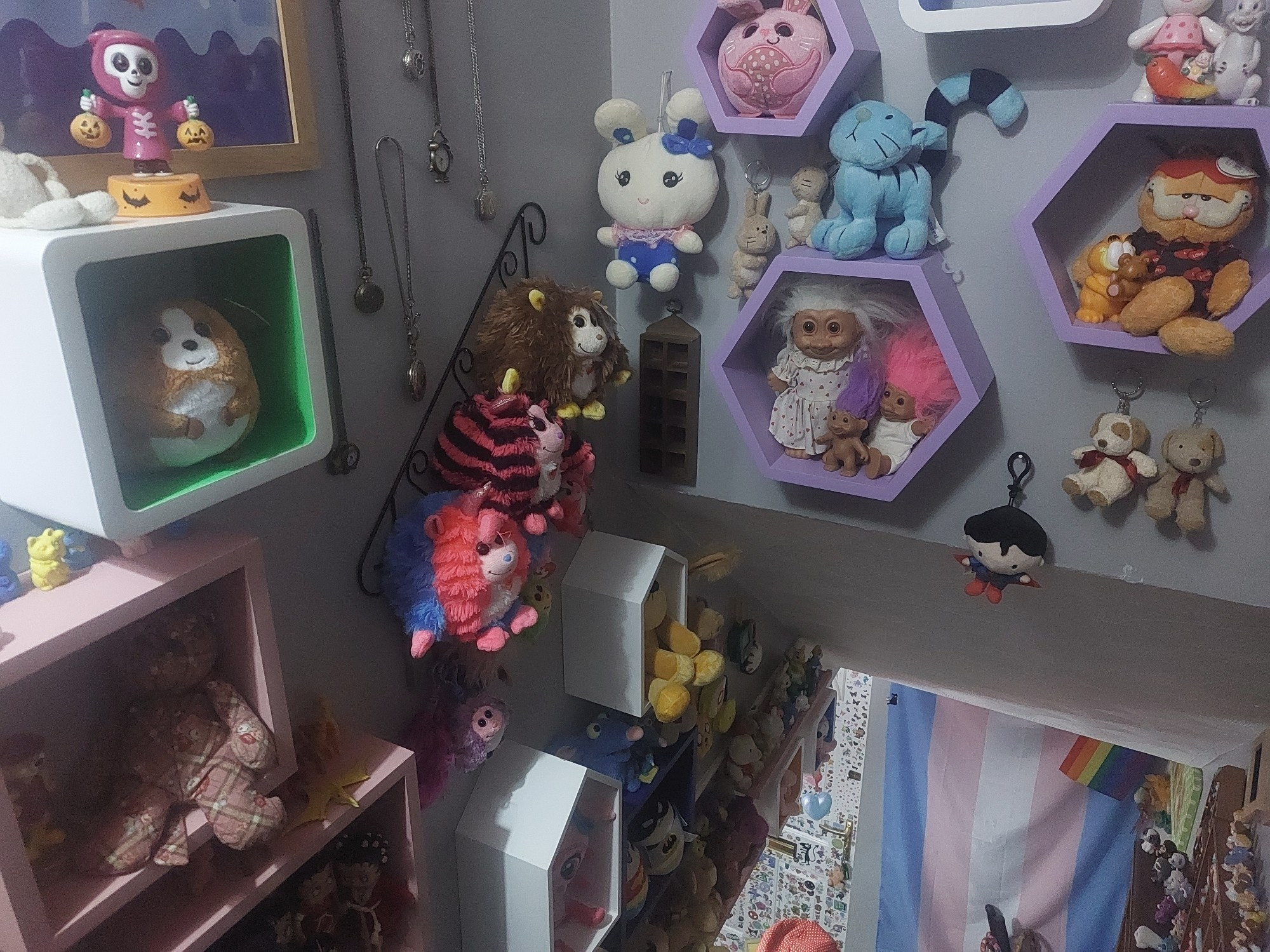 The view from the top of my stairs. Pride flags hang at the bottom, the front door is covered in stickers, and the walls are covered in shelves full of toys, keyrings and pocket watches.