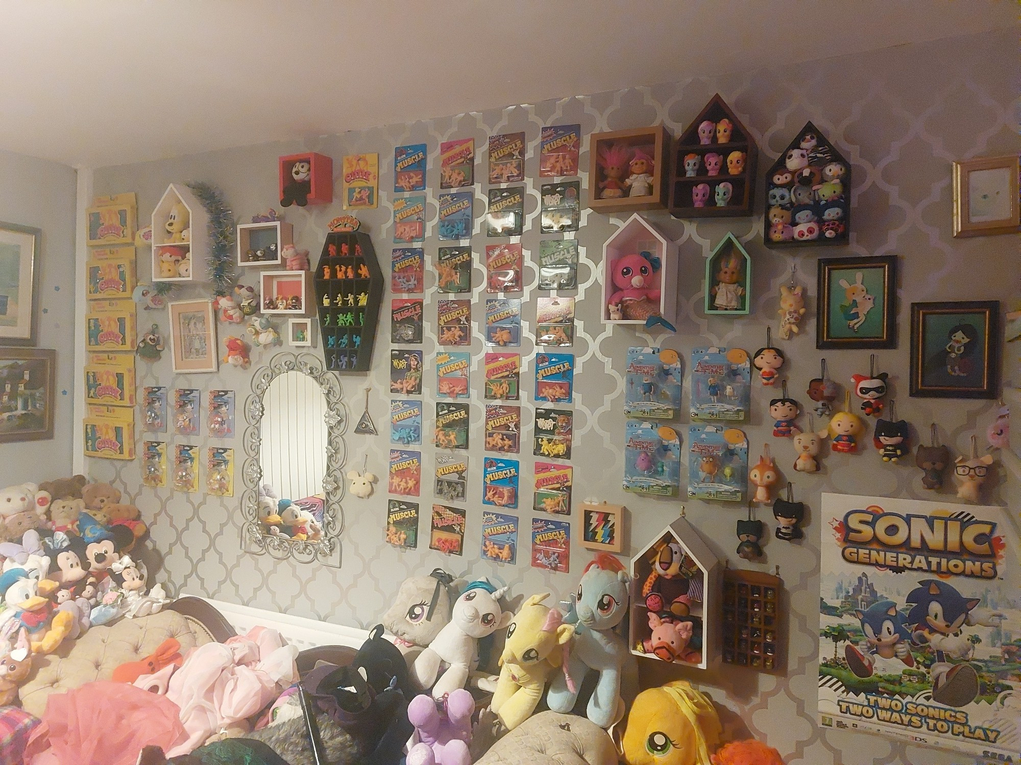 My living room wall. There's an old mirror, a coffin shaped shelf full of Monster in my Pockets arranged into a rainbow, a 4 x 8 grid of M.U.S.C.L.E. packs, some Adventure Time toys, more shelves of toys and figures, a Sonic poster, and a few other bits. In front of it is a sofa you can't currently sit on.