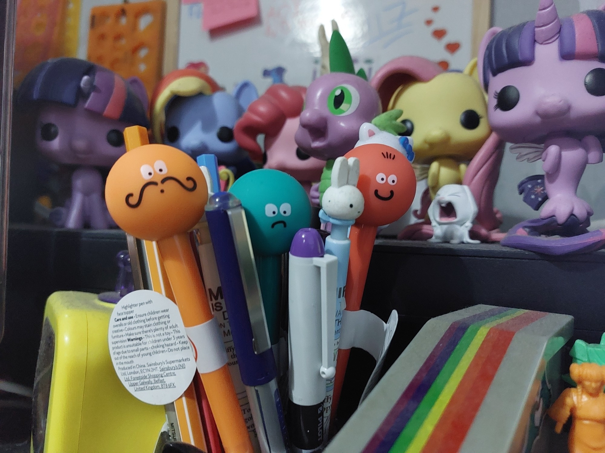 A close-up of my desk. There's pens (some novelty, some not), some Funko My Little Ponies, a TV shaped pet toy which I bought as a photo prop, and in the back a white board with stickers on and some drawing templates hanging from nails.