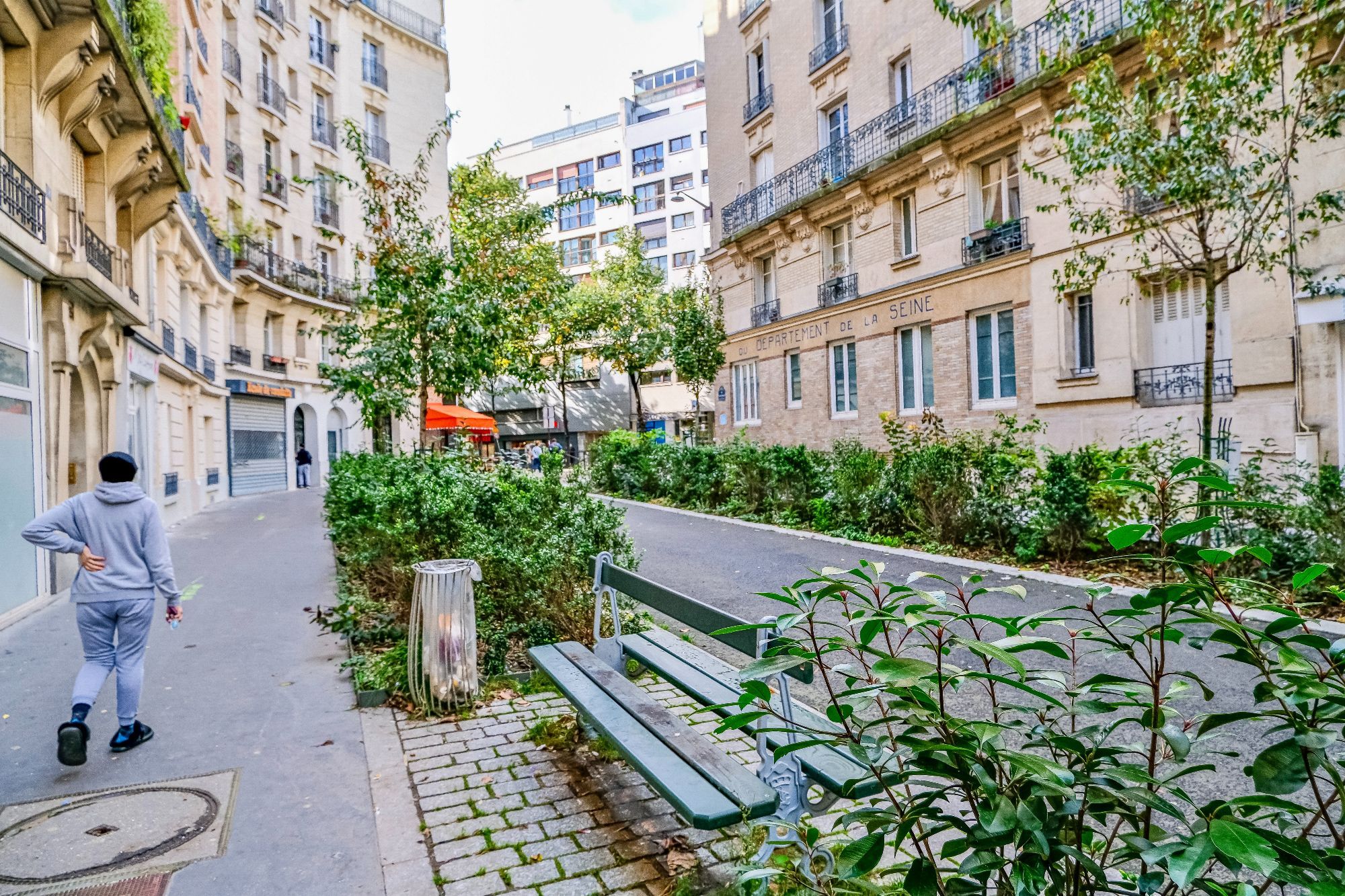 People walk and cycle through the 20th arrondissement of Paris. Rather than cars in the on-street parking spaces, they are filled with trees, terraces, benches, playgrounds, and parked bikes.