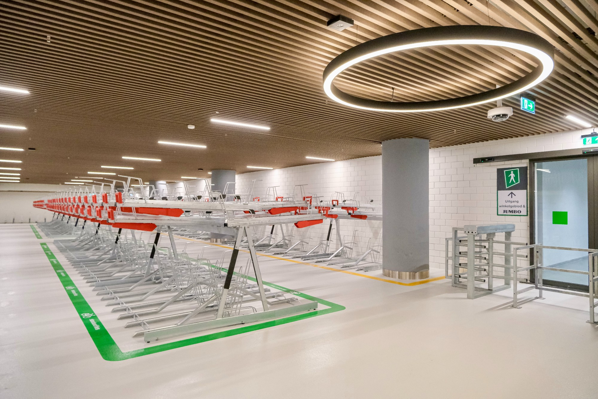 An underground bike parking facility in the city center of Rotterdam contains two-tiered metal racks, decorative lighting fixtures, and wood panelling on the ceiling.