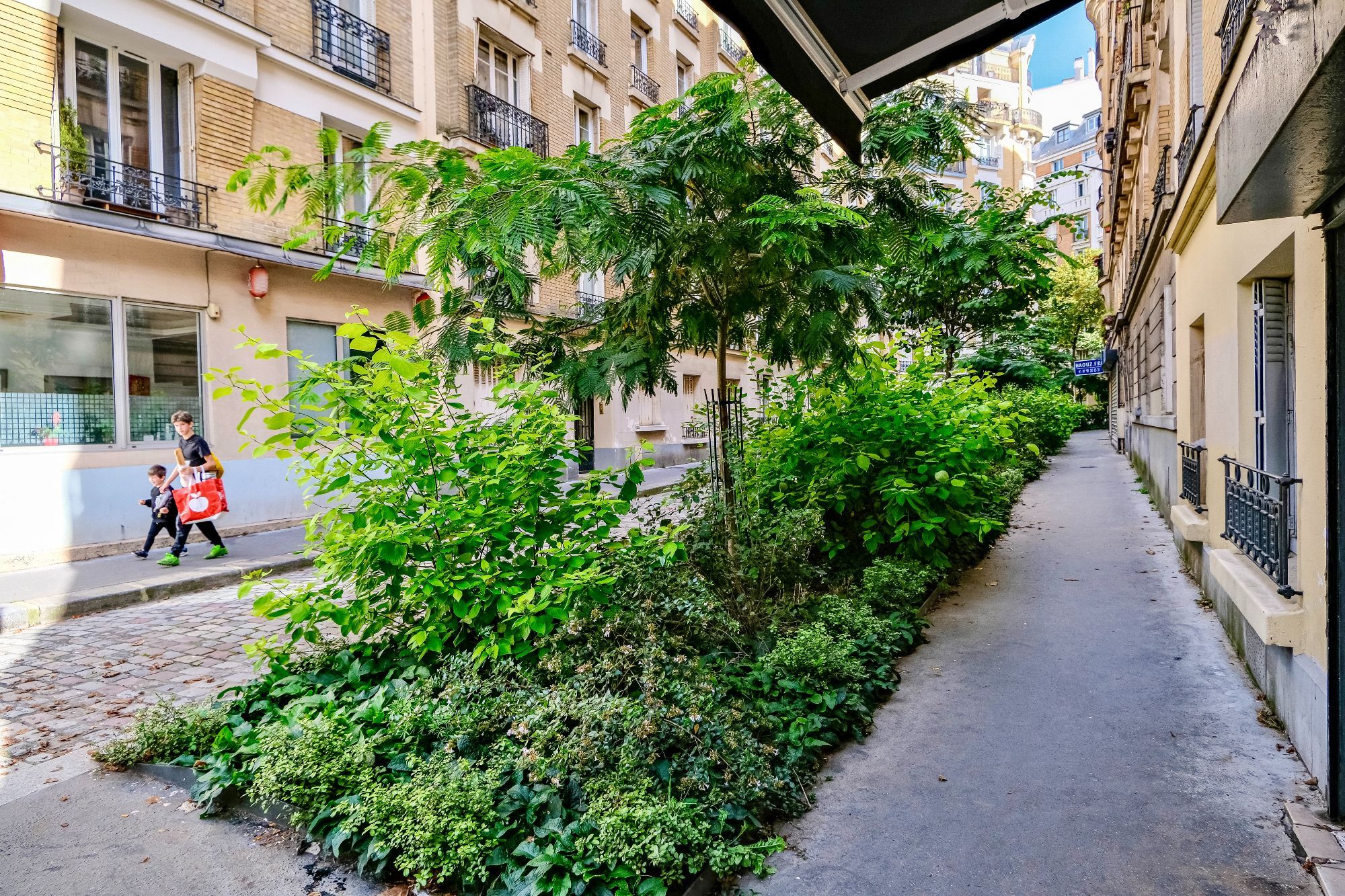 People walk and cycle through the 20th arrondissement of Paris. Rather than cars in the on-street parking spaces, they are filled with trees, terraces, benches, playgrounds, and parked bikes.