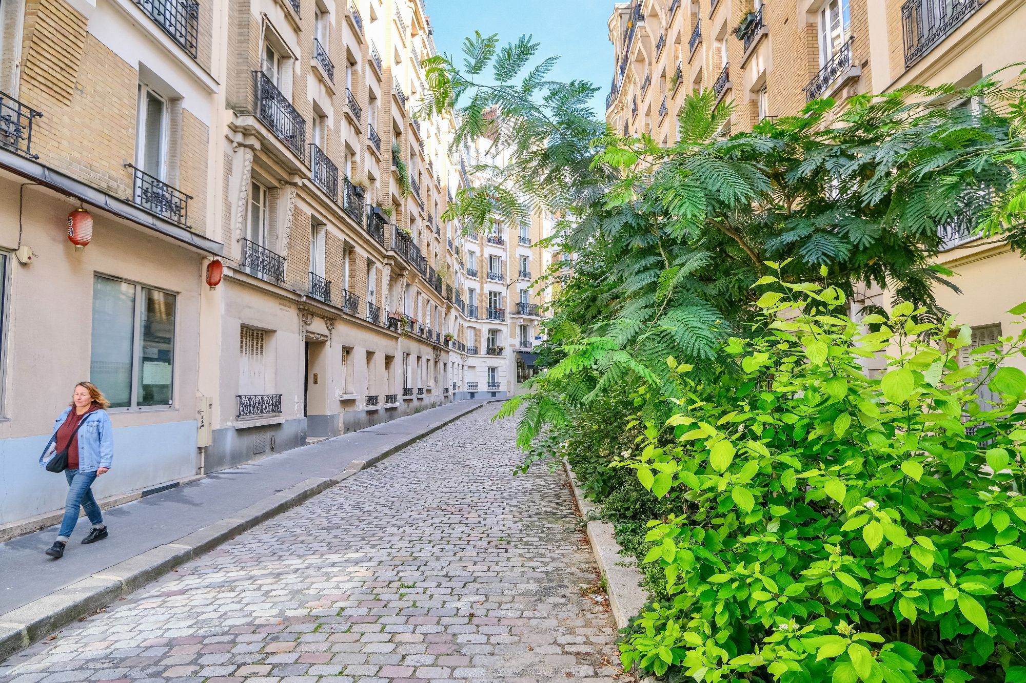 People walk and cycle through the 20th arrondissement of Paris. Rather than cars in the on-street parking spaces, they are filled with trees, terraces, benches, playgrounds, and parked bikes.