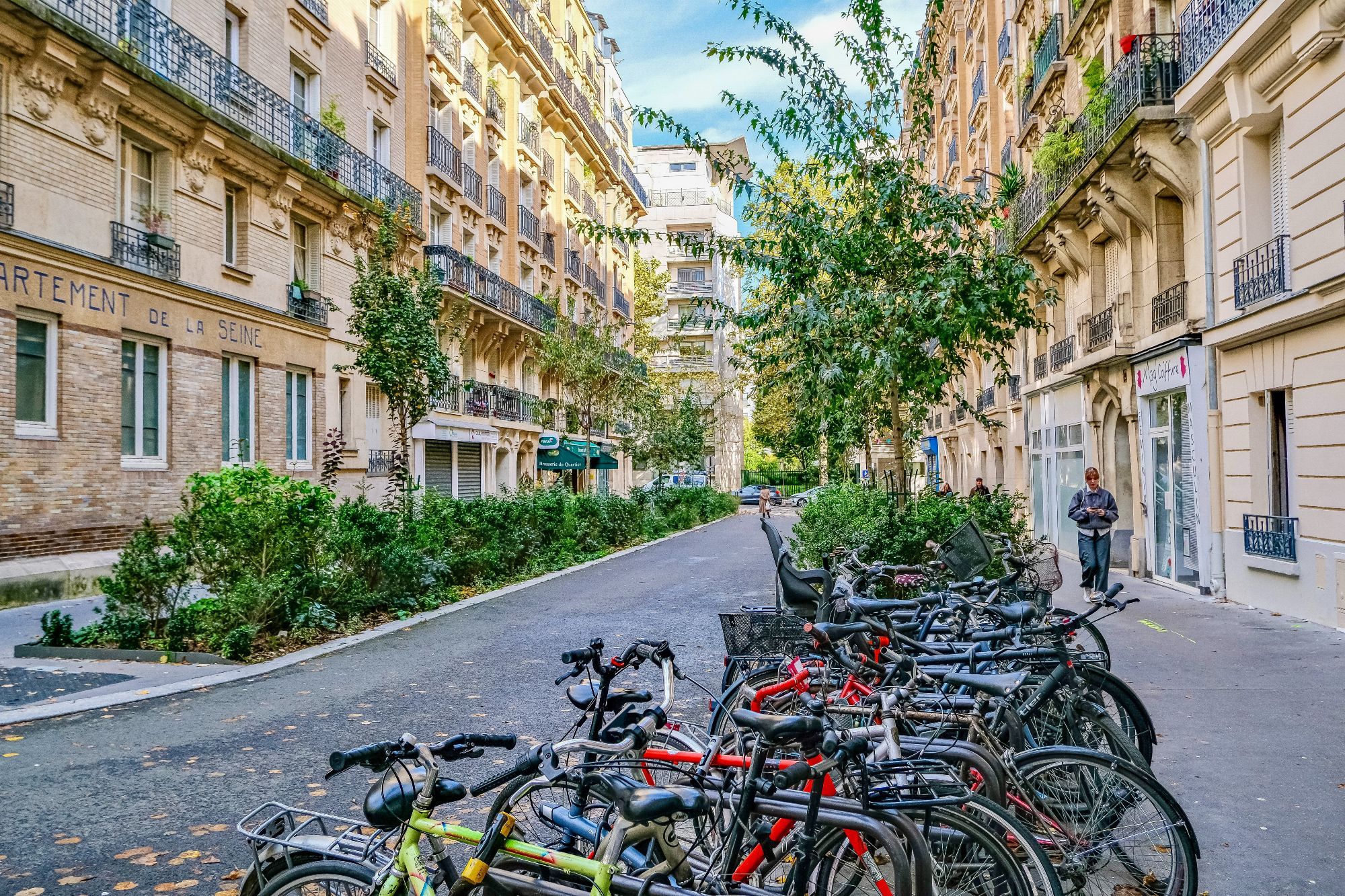 People walk and cycle through the 20th arrondissement of Paris. Rather than cars in the on-street parking spaces, they are filled with trees, terraces, benches, playgrounds, and parked bikes.