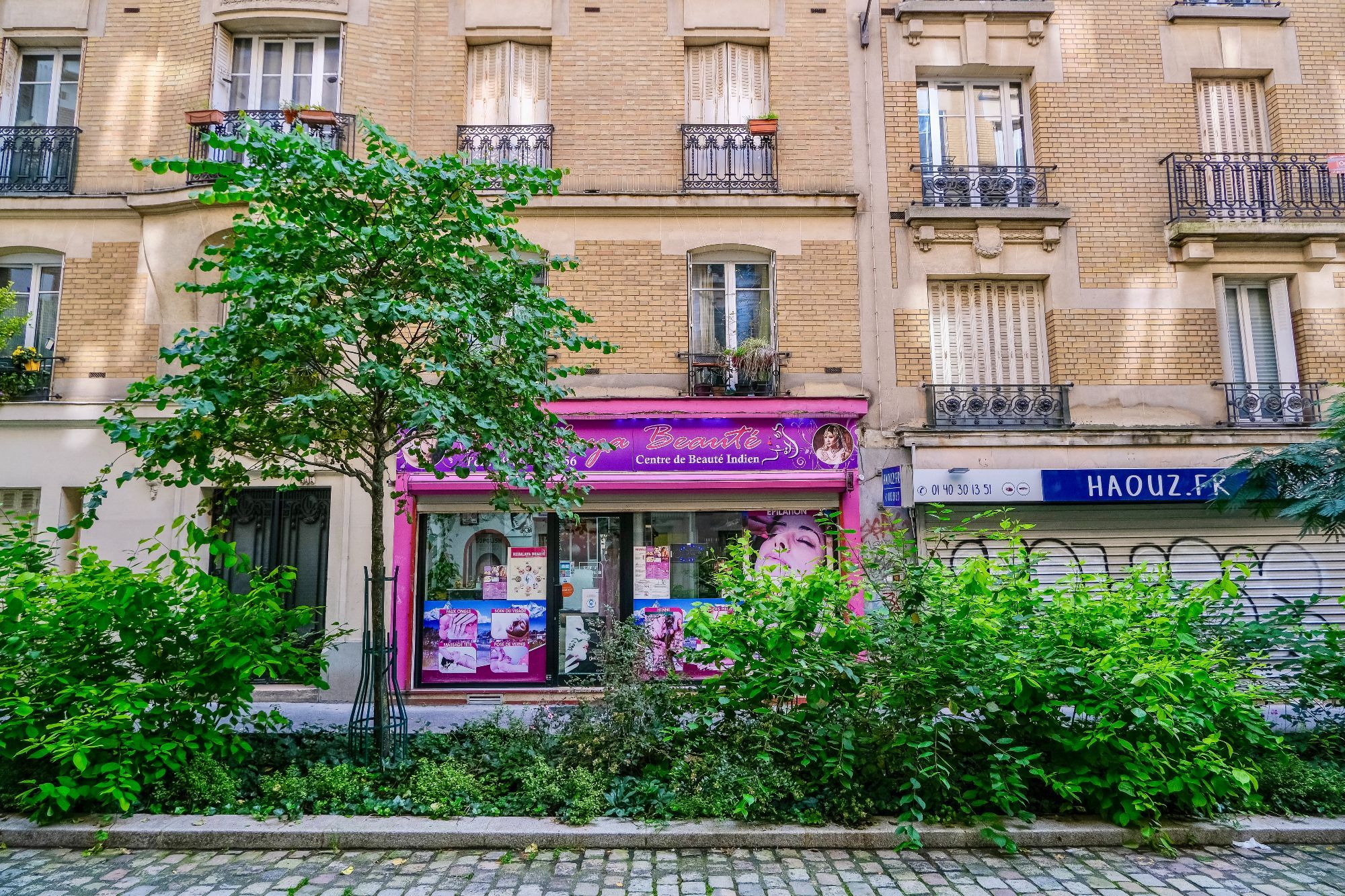 People walk and cycle through the 20th arrondissement of Paris. Rather than cars in the on-street parking spaces, they are filled with trees, terraces, benches, playgrounds, and parked bikes.