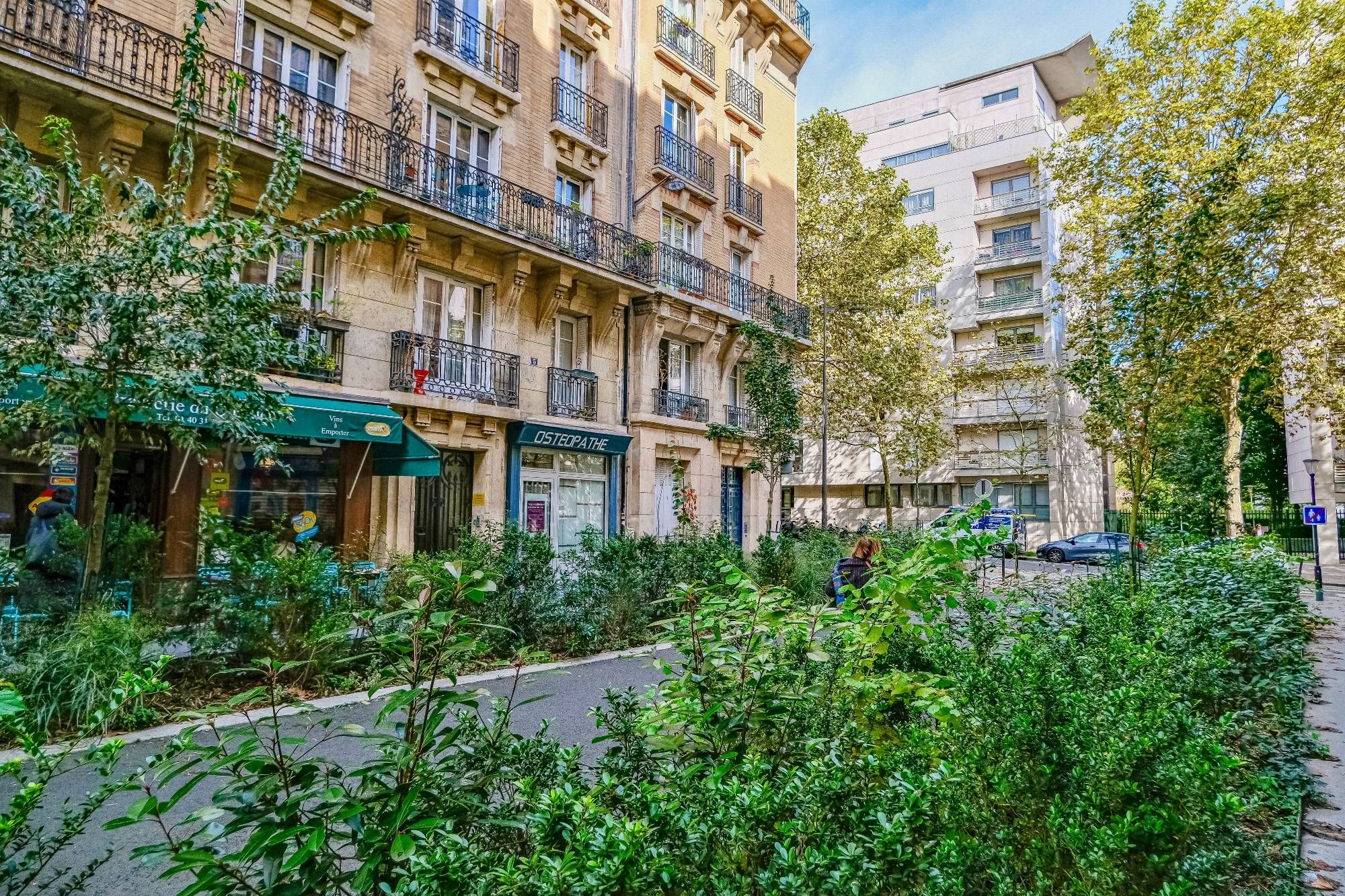 People walk and cycle through the 20th arrondissement of Paris. Rather than cars in the on-street parking spaces, they are filled with trees, terraces, benches, playgrounds, and parked bikes.