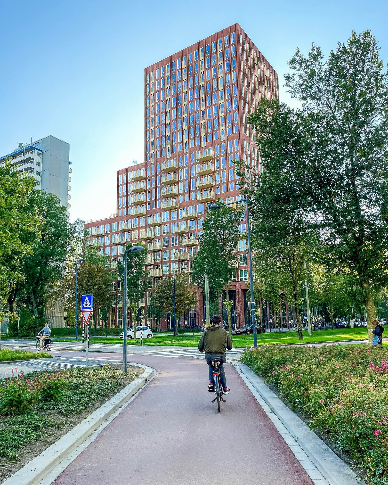 A teenage boy in casual attire rides along a red asphalt cycle path lined with trees, flowers, and tall apartment buildings in Delft.