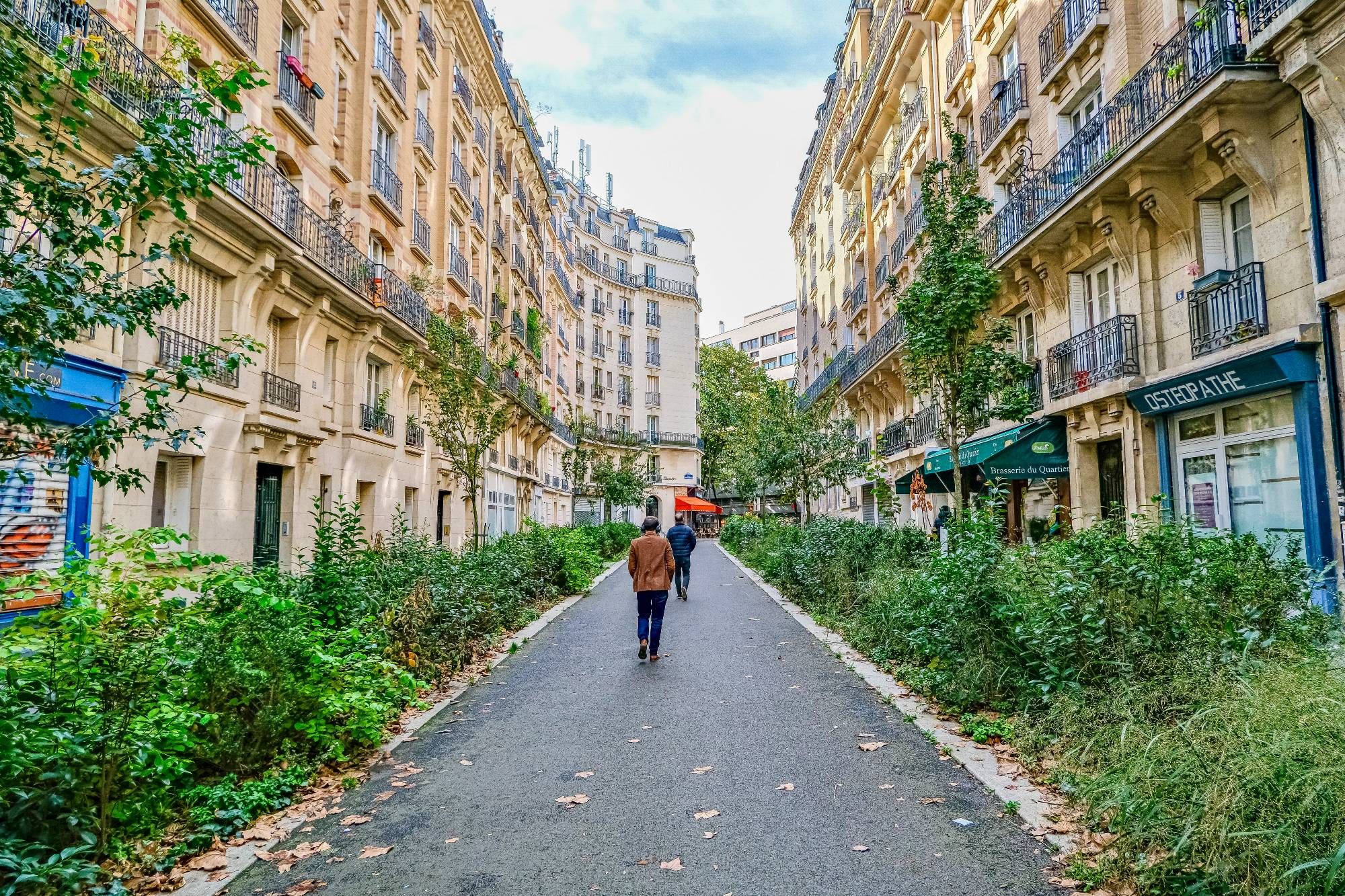 People walk and cycle through the 20th arrondissement of Paris. Rather than cars in the on-street parking spaces, they are filled with trees, terraces, benches, playgrounds, and parked bikes.