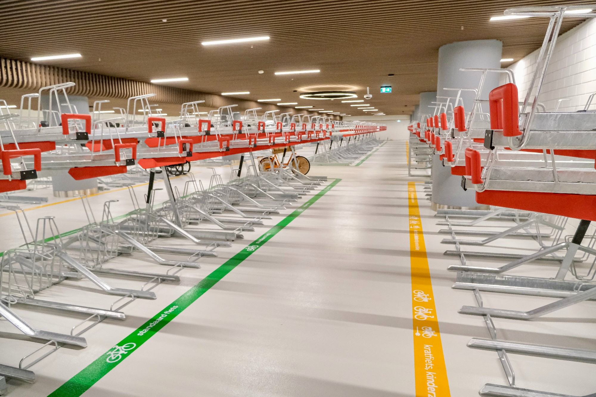 An underground bike parking facility in the city center of Rotterdam contains two-tiered metal racks, decorative lighting fixtures, and wood panelling on the ceiling.