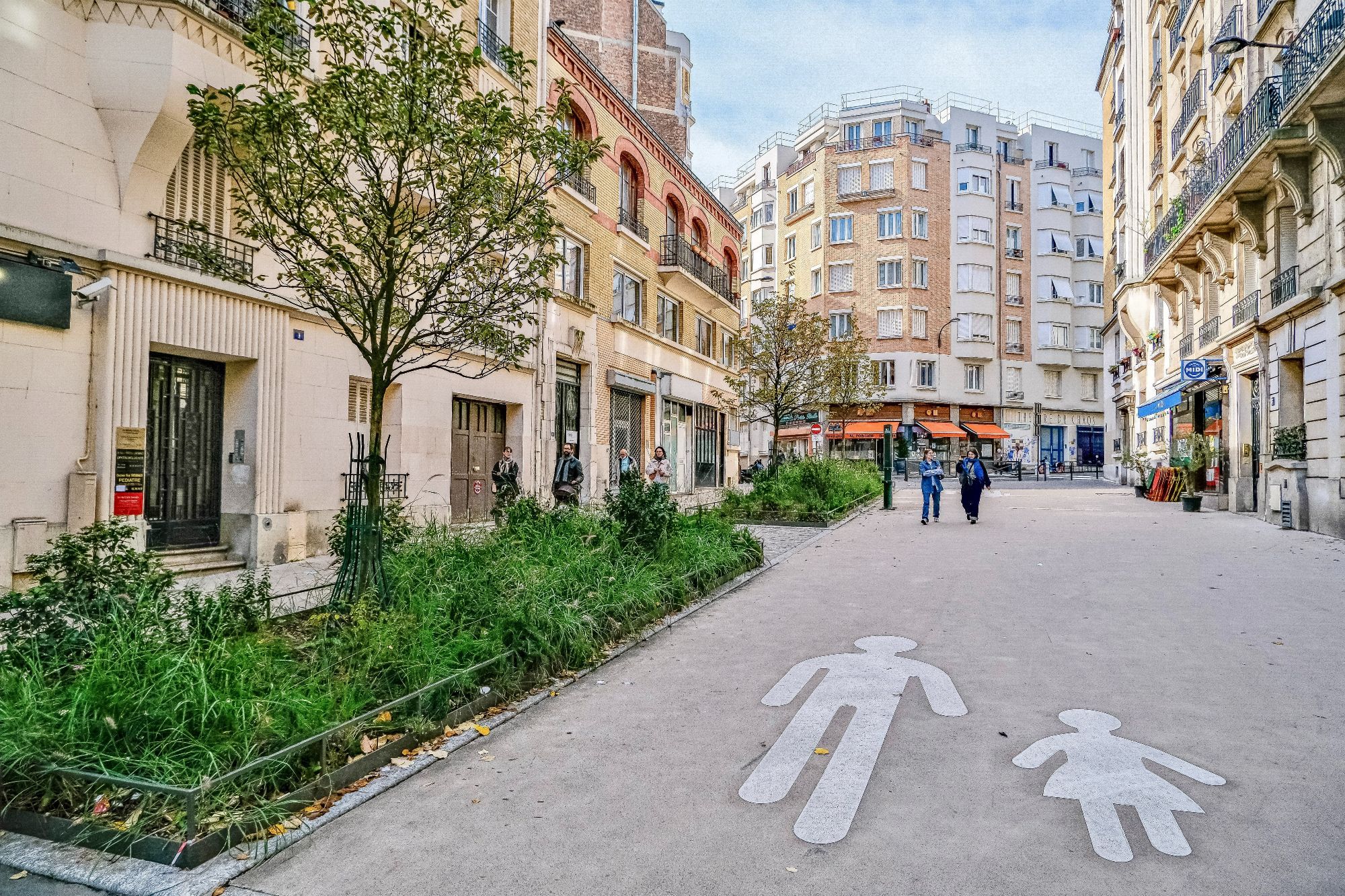 People walk and cycle through the 20th arrondissement of Paris. Rather than cars in the on-street parking spaces, they are filled with trees, terraces, benches, playgrounds, and parked bikes.