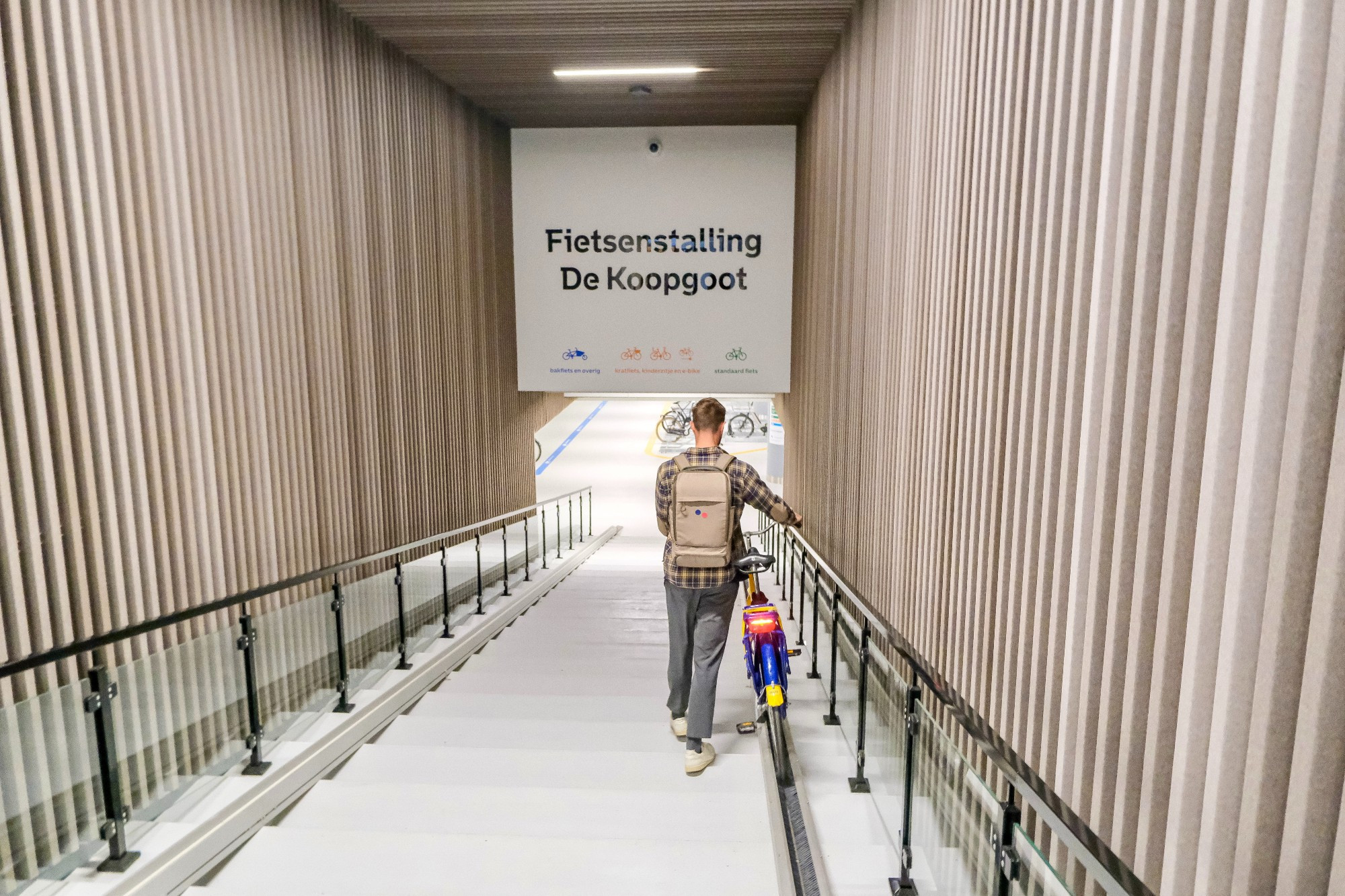 An underground bike parking facility in the city center of Rotterdam contains two-tiered metal racks, decorative lighting fixtures, and wood panelling on the ceiling.