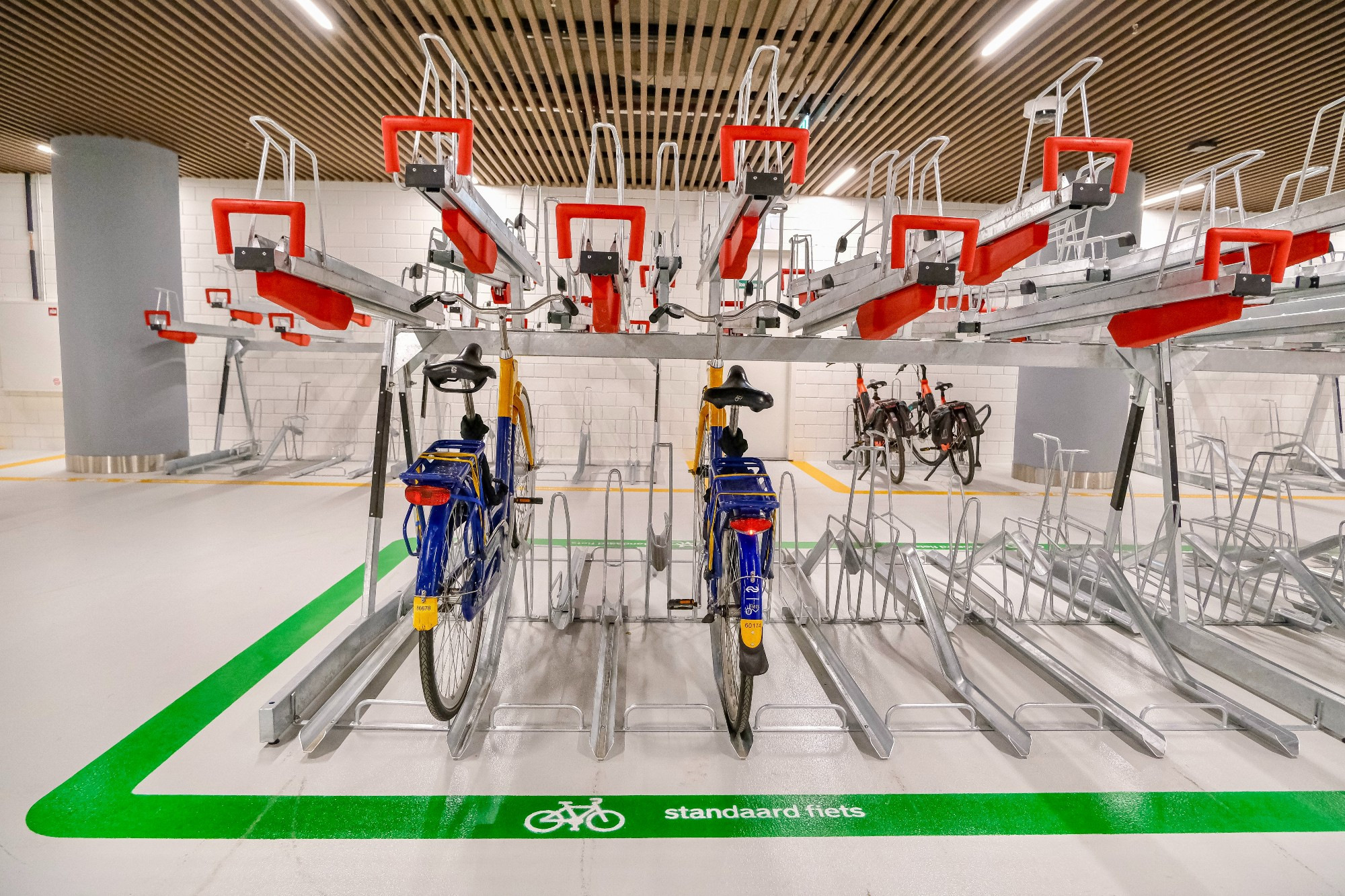 An underground bike parking facility in the city center of Rotterdam contains two-tiered metal racks, decorative lighting fixtures, and wood panelling on the ceiling.