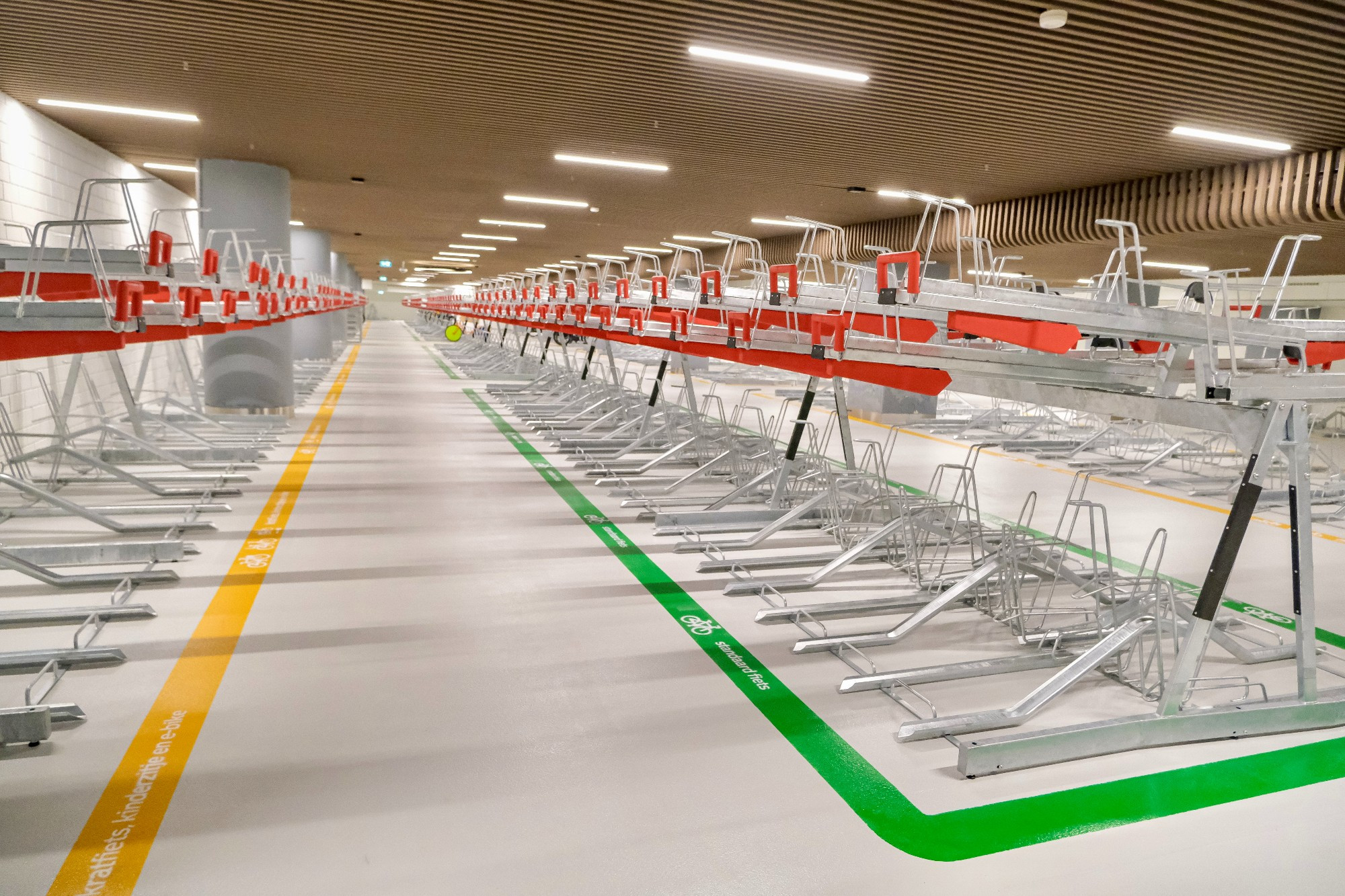 An underground bike parking facility in the city center of Rotterdam contains two-tiered metal racks, decorative lighting fixtures, and wood panelling on the ceiling.