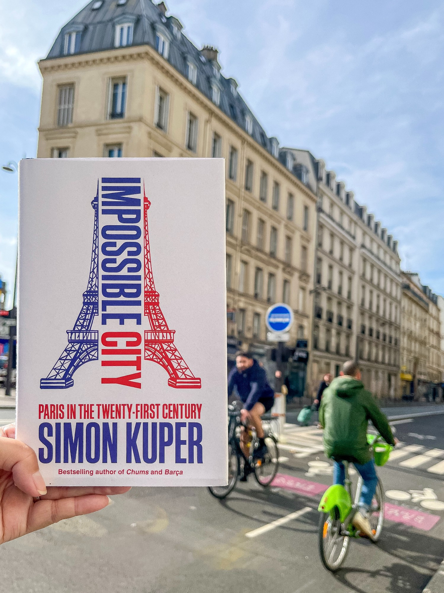 A hand holds a hardcover book: "Impossible City" by Simon Kuper, in front a building and bike lane in the center of Paris.