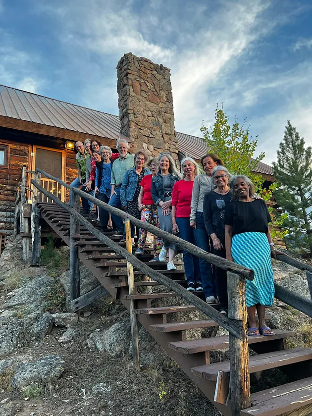 Thirteen men and women writers, participants in a writing retreat, standing on steps leading from a rustic log cabin chapel on a cliffside in the mountains. A blue sky with white clouds and pine and aspen trees also in the photo. 