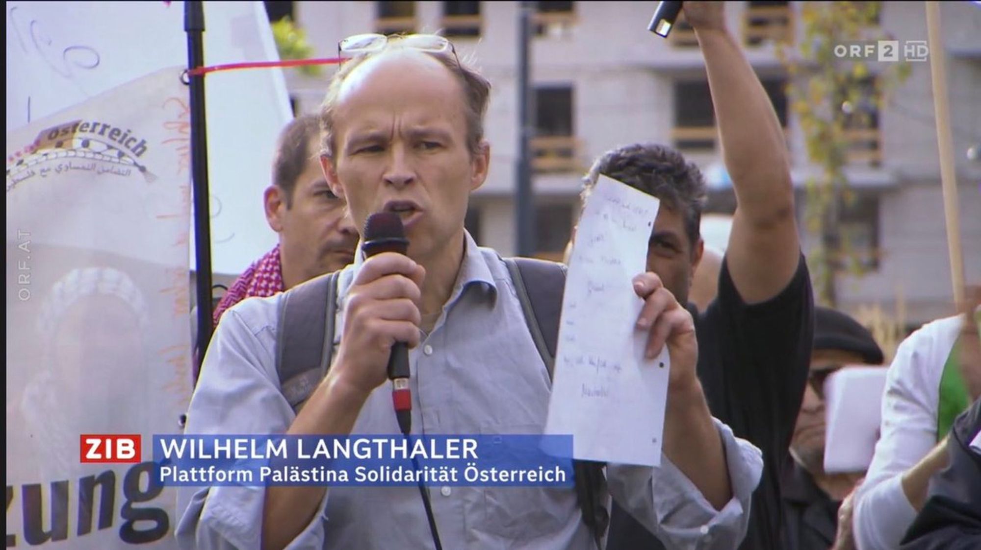 Screenshot aus der Zeit im Bild, man sieht Wilhelm Langthaler auf einer antiisraelischen Kundgebung am Columbusplatz, Wien, mit einem Mikrofon in seiner Hand, dahinter Martin Weinberger mit roter Kufiya, er ist ebenfalls bei Palästinasolidarität aktiv.