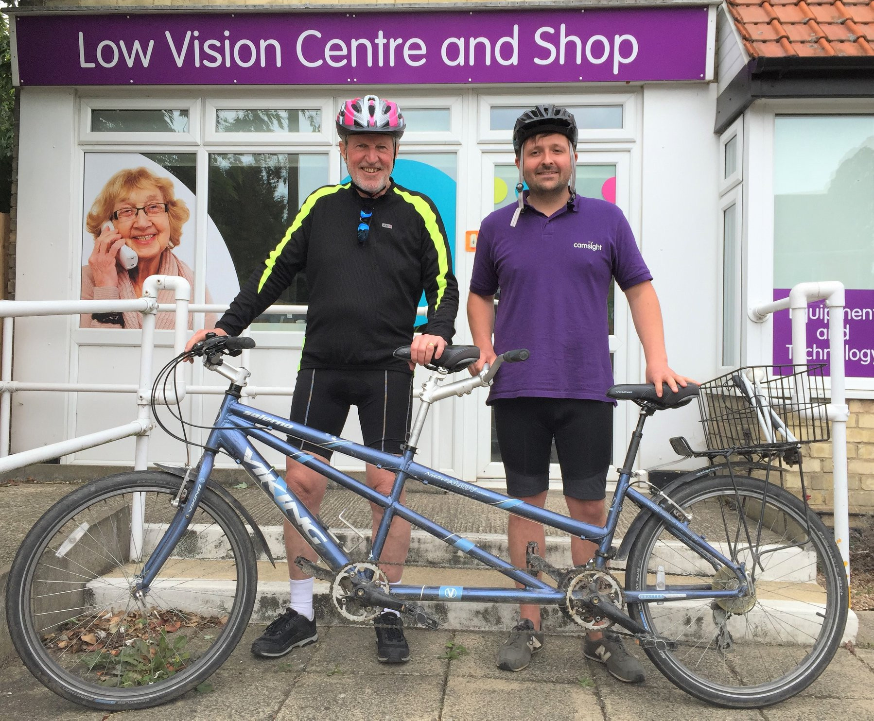 A sighted pilot and stoker with vision issues wearing a purple Cam Sight T-shirt stand behind a blue Viking tandem outside the Cam Sight Low Vision Centre and Shop. Both are wearing cycling shorts and helmets.