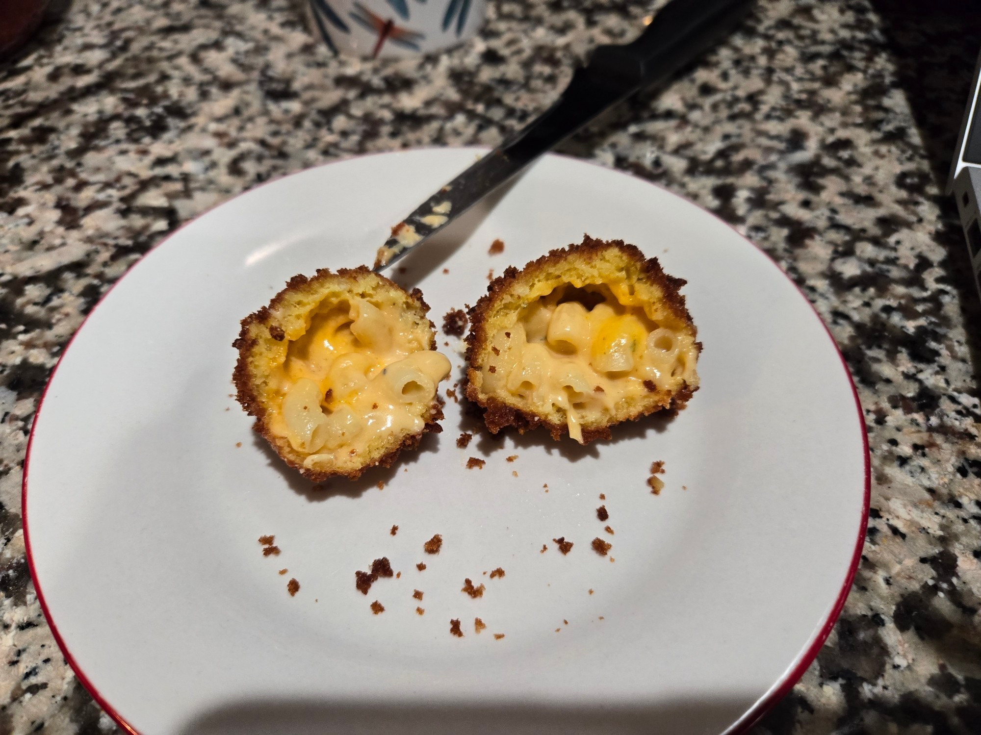 Two halves of a fresh, 5 cheese, fried Mac & Cheese ball on a saucer with a knife above.