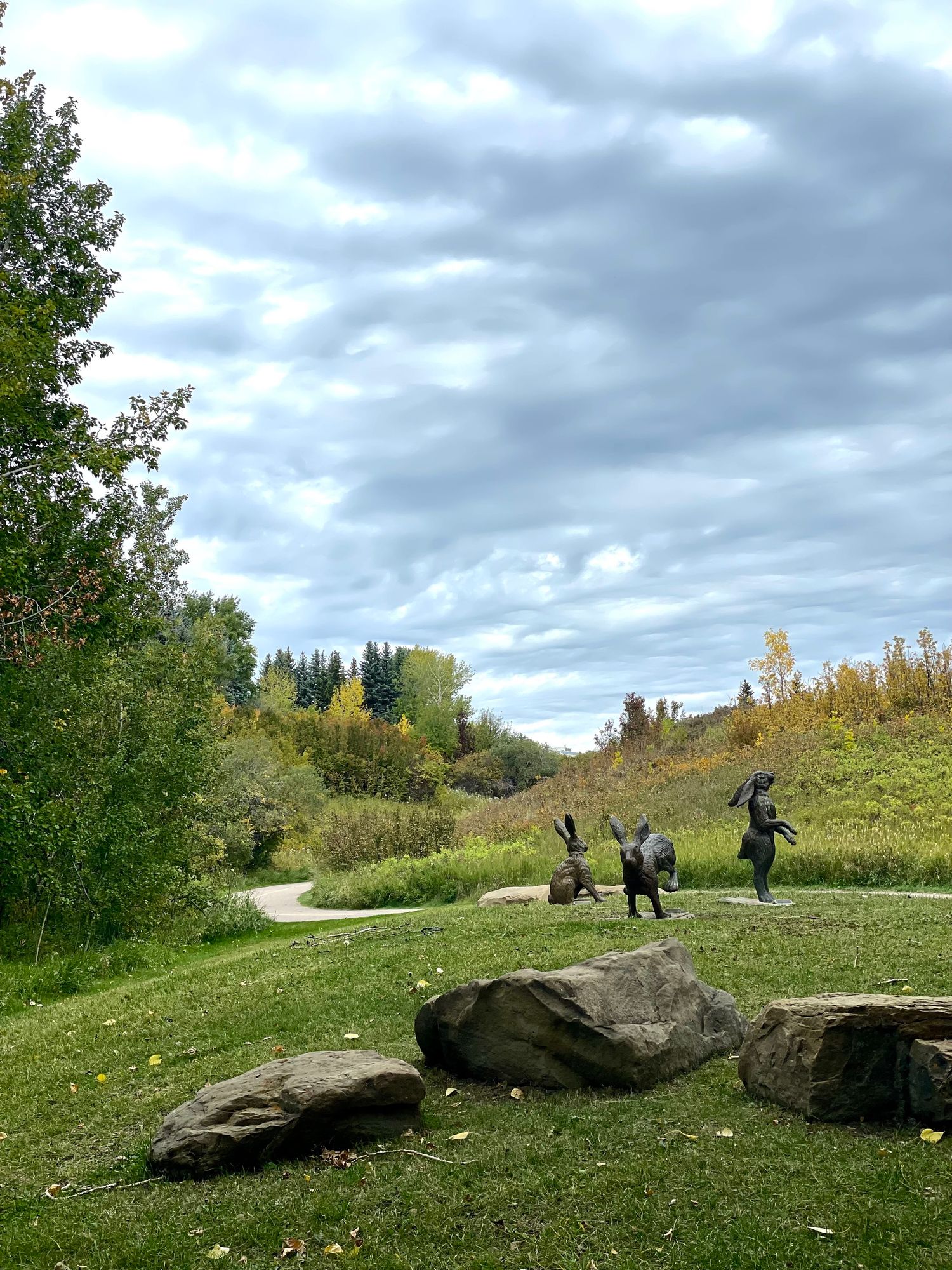 A grey, cloudy autumn sky behind rolling hills. The leaves have started to turn yellow mixed with green. Three bunny statues are arranged in a loose circle by a pathway. One sitting, one running, and one standing to look at the sky. In the immediate foreground are three large, flattish rocks.