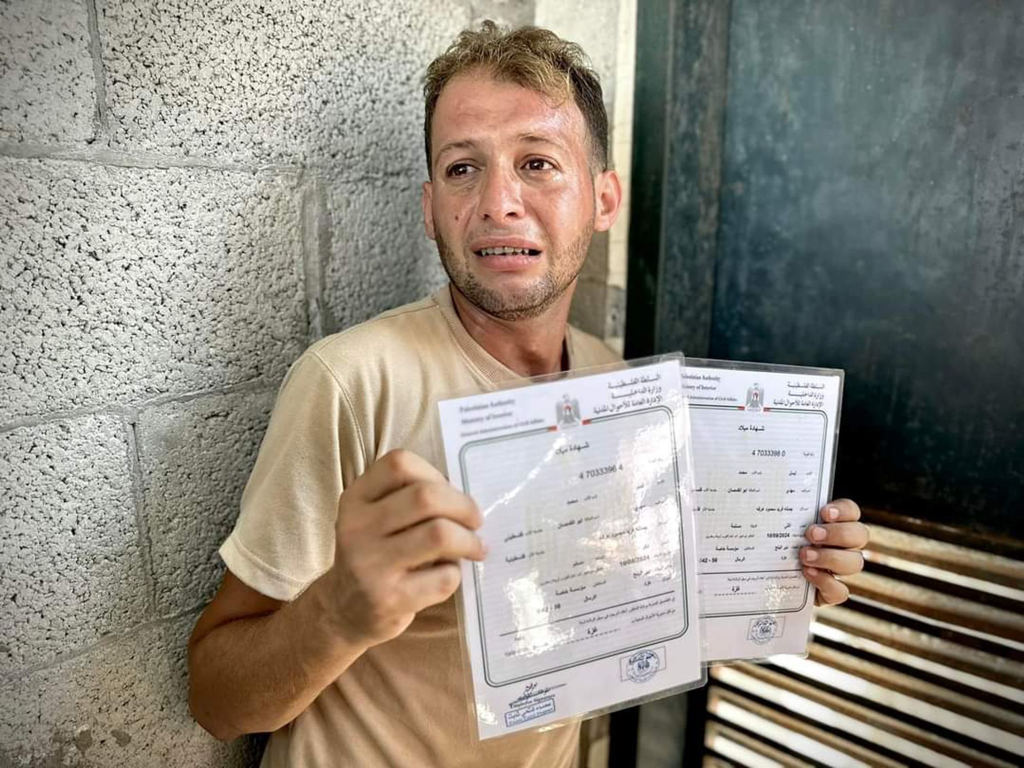 Father of 4-day-old twins, Mohammad Abu al-Qumsan, holds up two laminated birth certificates. He is wearing a beige t-shirt and sitting in front of a concrete block wall. He has been weeping.