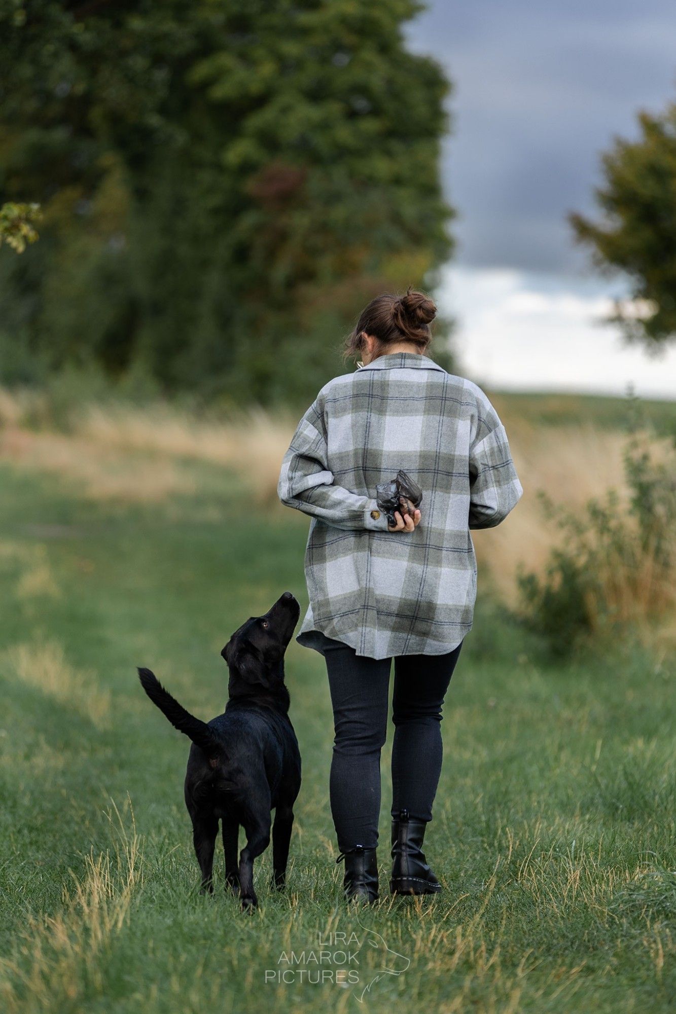 Eine Frau in schwarzer Jeans und grau-grünem Flanellhemd läuft über einen Feldweg von der Kamera weg. Sie hält eine Hand auf dem Rücken, in der sie einen Kackbeutel, der mit Käse und Schinken gefüllt ist. Neben ihr läuft ein schwarzer Labrador, der sie ganz aufmerksam ansieht. Das Foto symbolisiert die Bindung zwischen den Beiden.