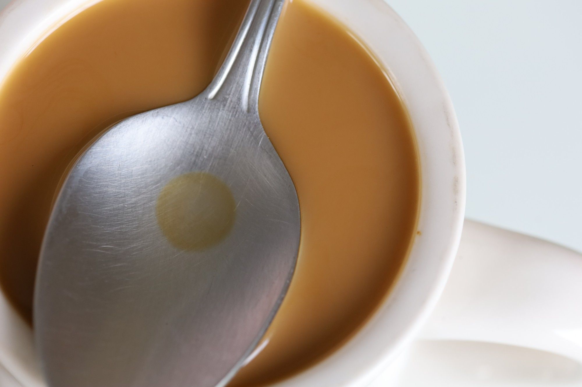 Macro photo of a thick white mug of light brown coffee. On top of the mug is a stainless steel spoon with a dried coffee "dot".