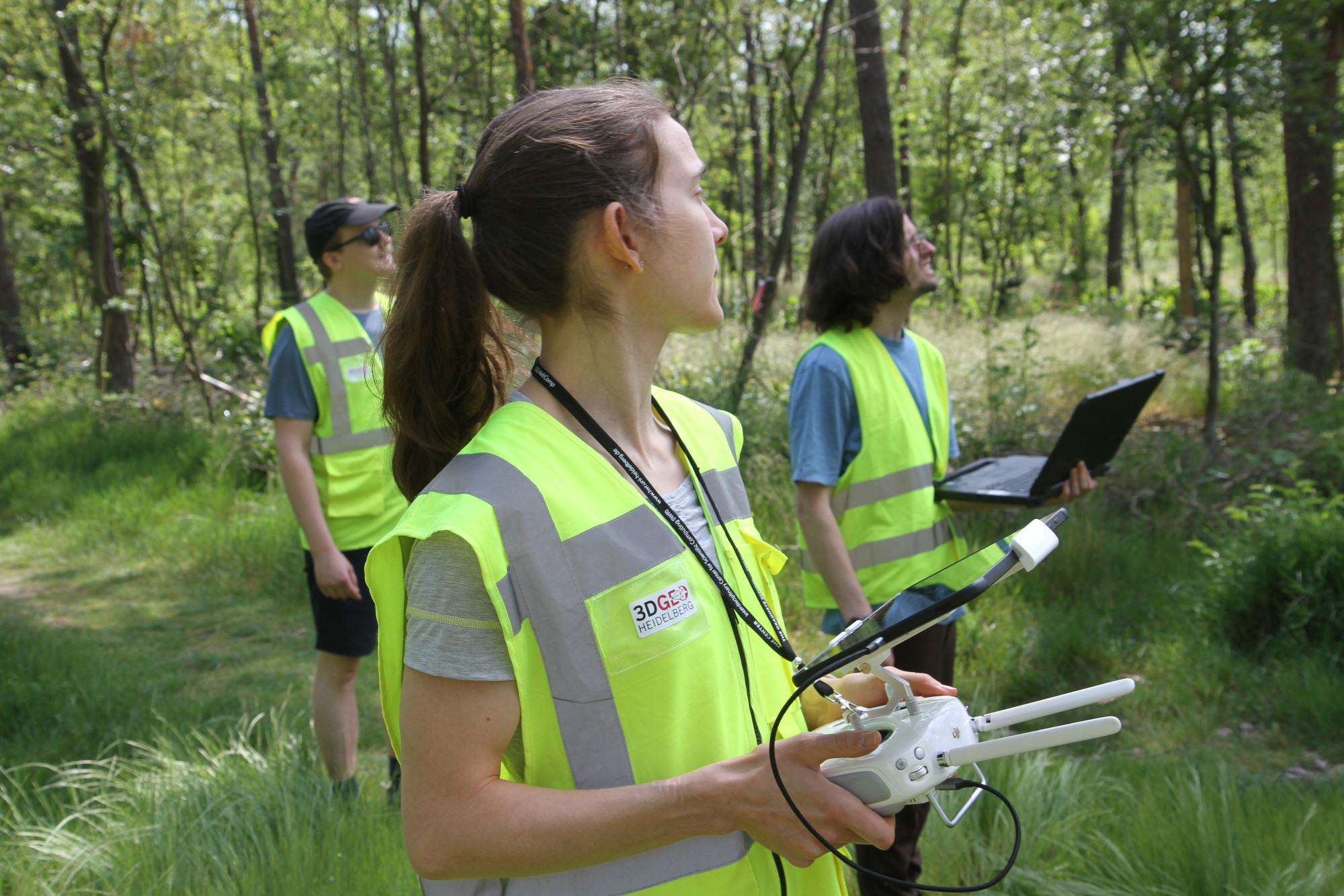 Three people in yellow warn wests looking at the sky in the same direction. The person in the front carries the remote control of a UAV, another person carries a field laptop.