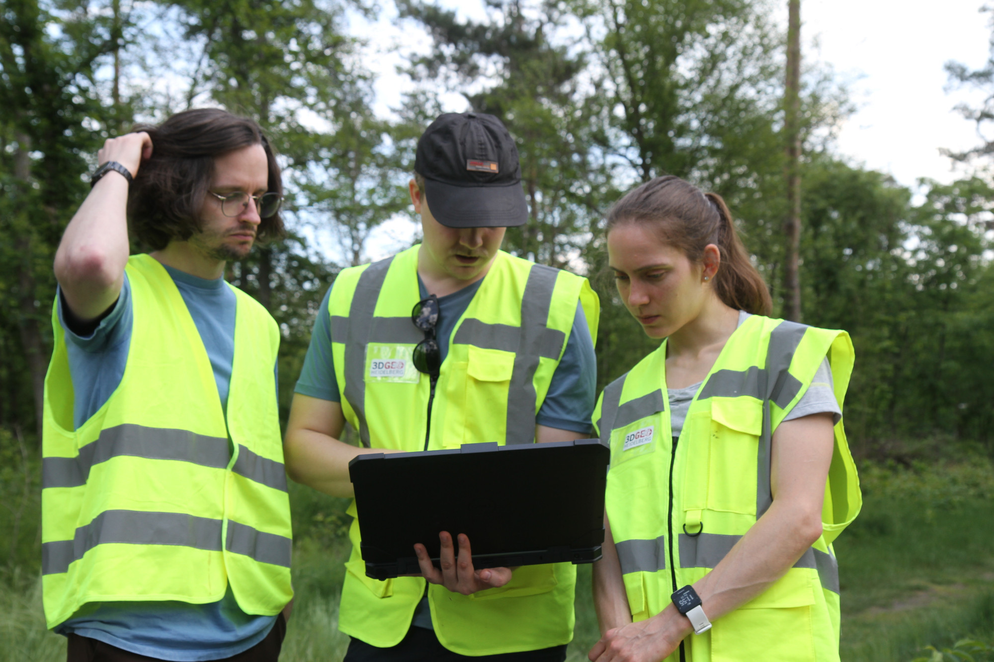 Three people in warn wests (with a 3DGeo Heidelberg Logo in) are looking at the screen of their field laptop.