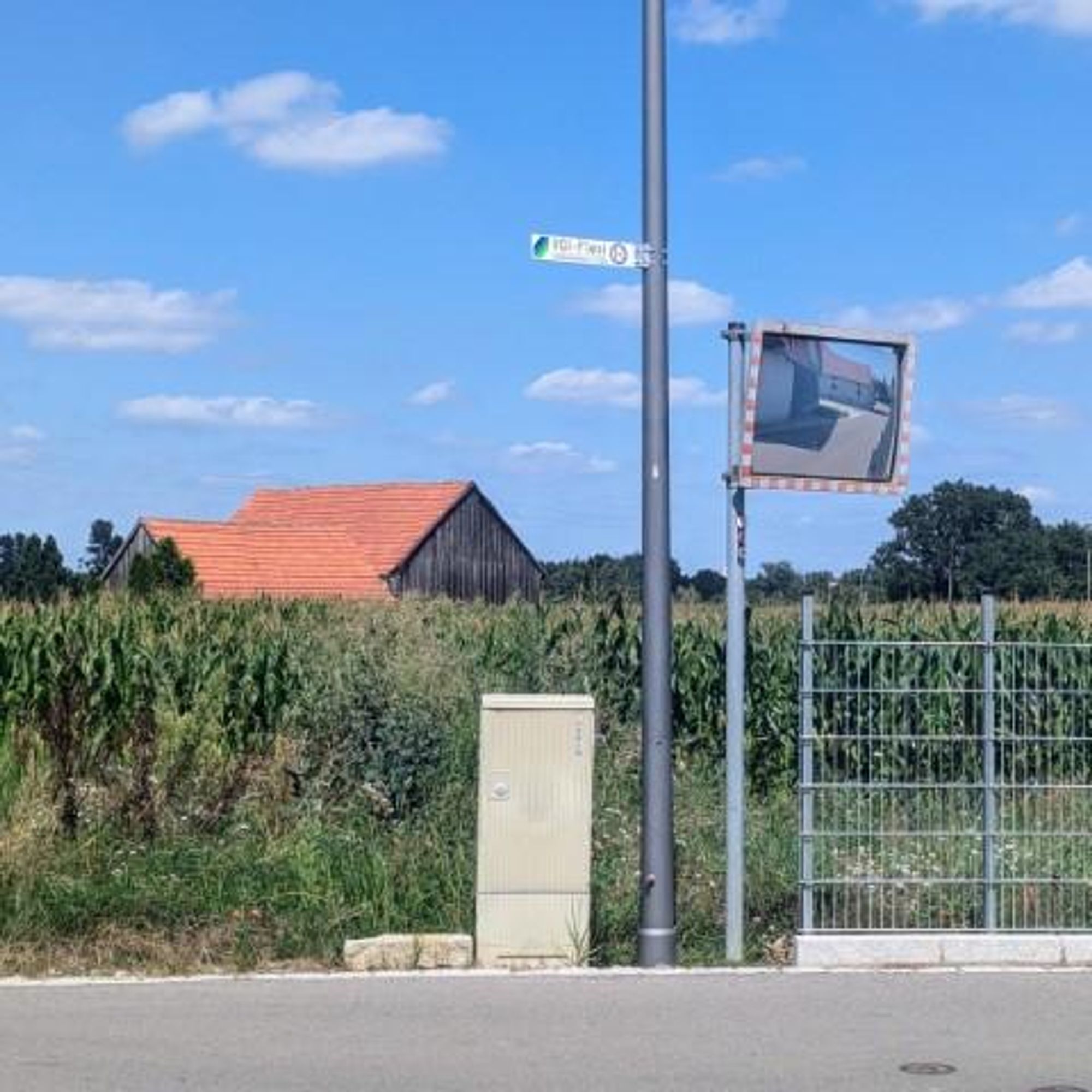 Das Bild zeigt eine ländliche Straßenszene unter einem klaren blauen Himmel. Links steht ein Laternenmast mit einem grünen Straßenschild "VGI Flexi". Neben dem Laternenmast ist ein rechteckiger Verkehrsspiegel angebracht, in dem sich die benachbarten Gebäude spiegeln. Hinter den Laternenmast befindet sich ein Maisfeld. Davor verläuft die Kramerstraße in Mändlfeld.