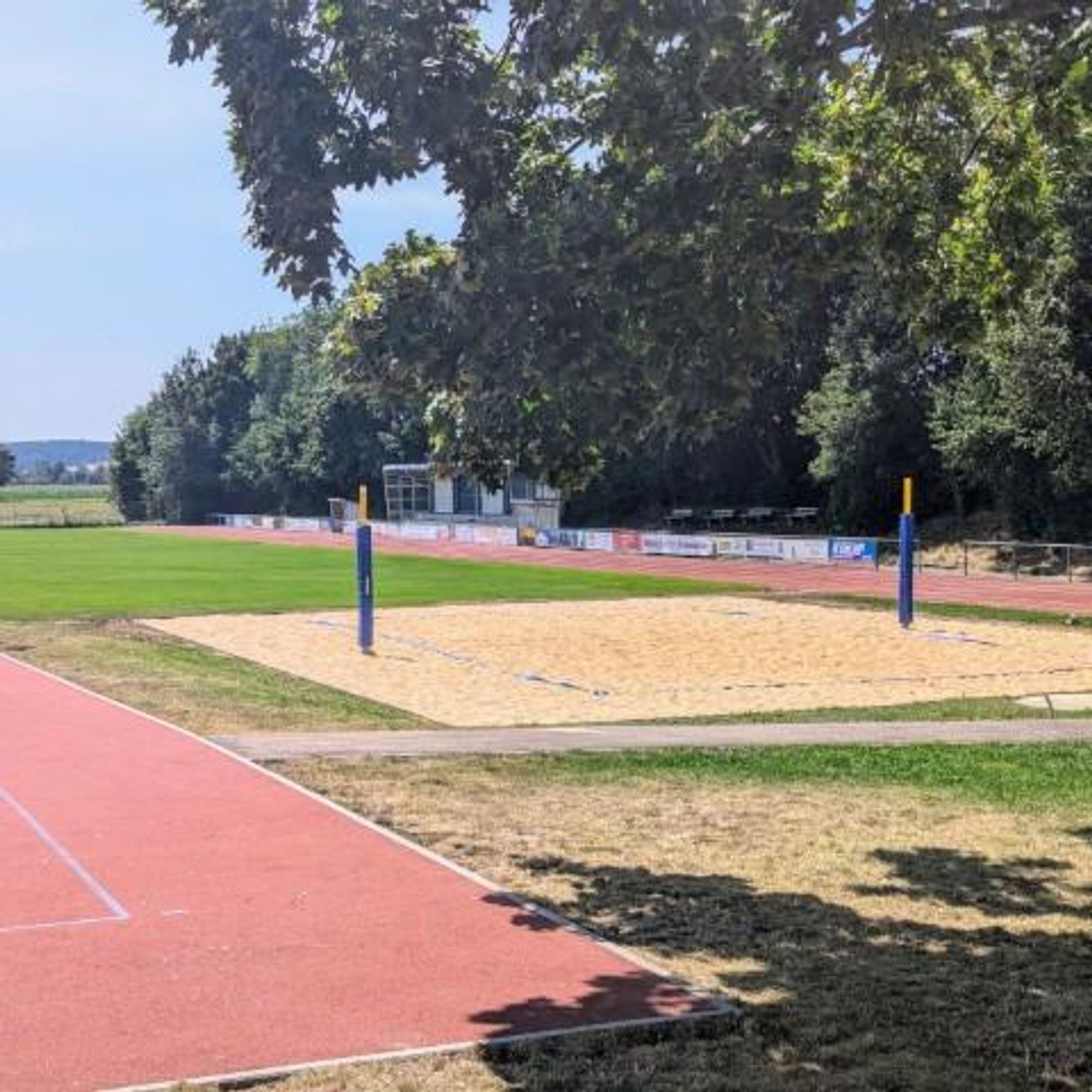 Der Sportplatz Karlskron an einem sonnigen Tag. Auf der rechten Seite befindet sich eine rote Laufbahn. Links ist eine Ecke des rotten Hartgummiplatzes sichtbar. Dazwischen liegt das neu geschaffene Beachvolleyballfeld mit goldgelben Sand. Das Feld ist durch blaue Kunststoffbänder gekennzeichnet und die Pfosten für das Netz mit blauem Schaumstoff eingehüllt. Das Netz ist nicht zu erkennen.