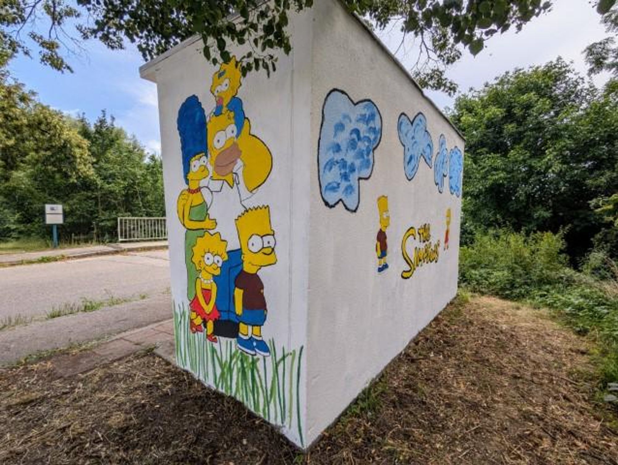 Das Bild zeigt eine Wandmalerei der animierten Fernsehserie “Die Simpsons”. Die Malerei befindet sich im Brautlacher Bushäuschen. Zu sehen sind die östliche Außenseite und die Rückseite des Bushäuschens. Die Rückseite zeigt den Simpsons-Schriftzug. Auf der Seitenwand ist die Familie Simpson zu sehen, die den Betrachter anlächelt.