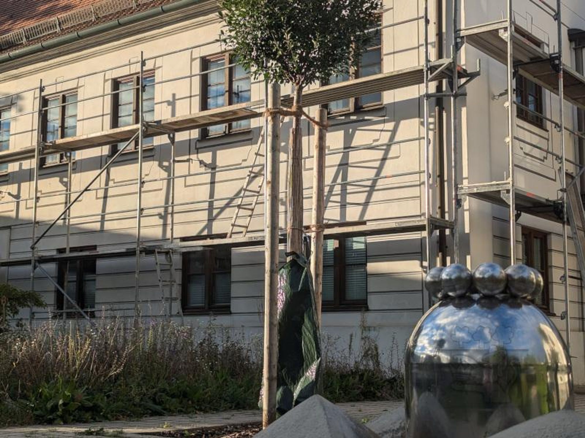 Das Bild zeigt das Rathaus von Karlskron, das derzeit von einem Baugerüst umgeben ist. Das Gebäude hat eine helle Fassade und mehrere Fenster. Das Gerüst besteht aus Metallstangen und umgibt das gesamte Gebäude. Im Vordergrund befindet ein kleiner Baum und der Gemeindebrunnen.