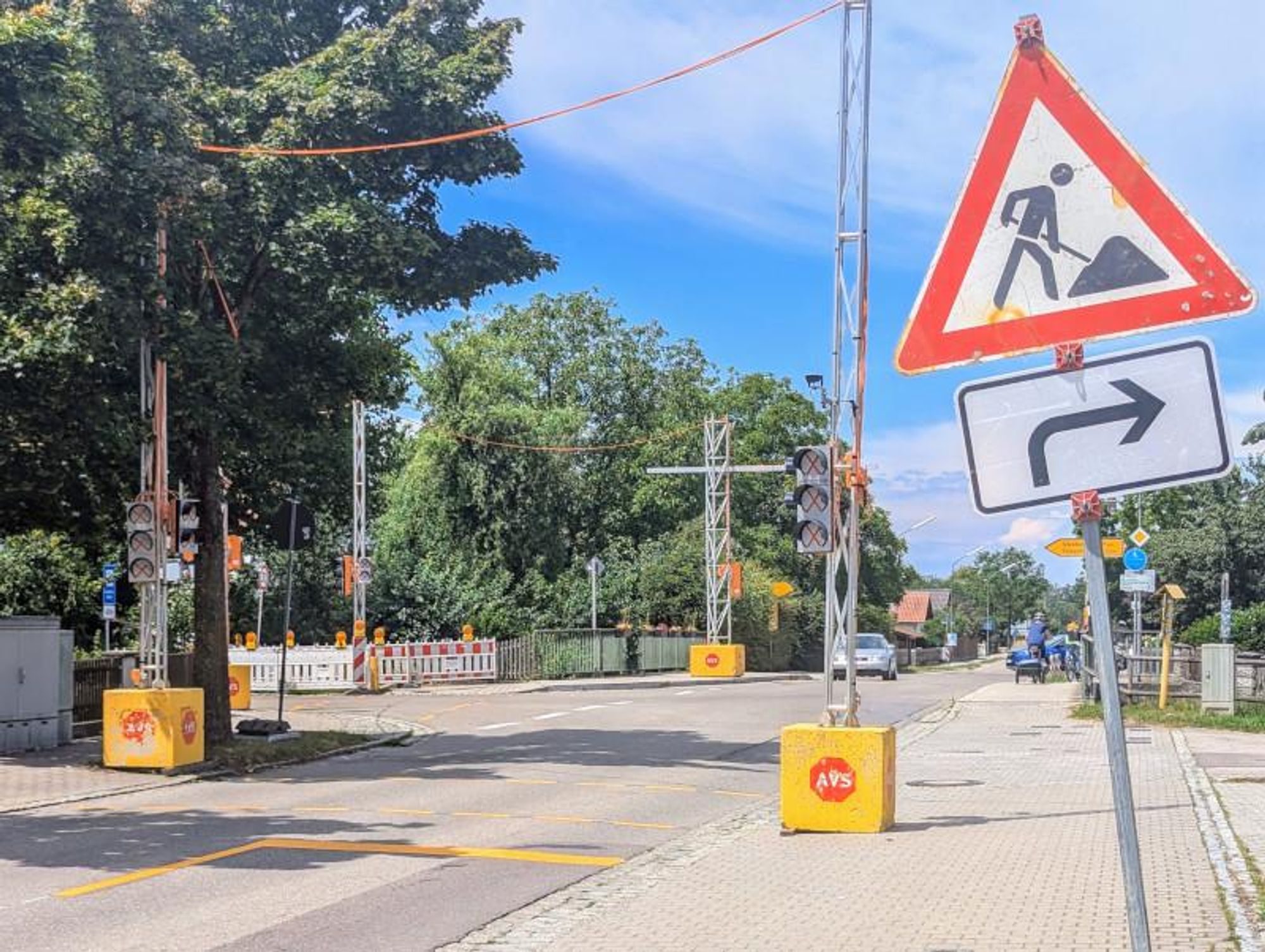 Ein Foto einer temporären Verkehrsampel an einer Straßenkreuzung. Die Ampel steht auf einem provisorischen Ständer und ist noch nicht in Betrieb. Die Lampen der Ampel sind abgedeckt. Im Hintergrund sind verschwommene Silhouetten von Gebäuden zu erkennen. Es scheint ein sonniger Tag zu sein, da der Himmel im Hintergrund blau ist.