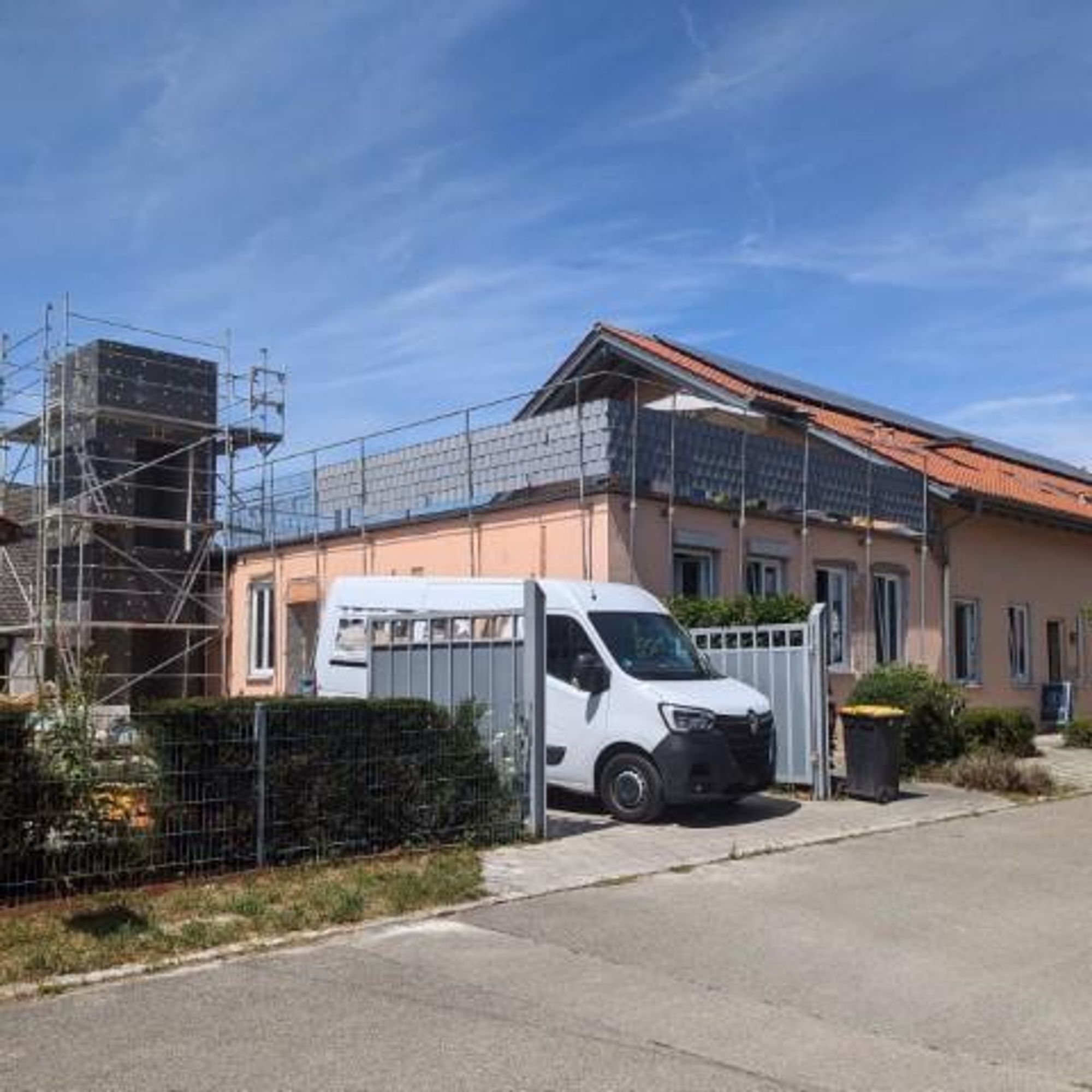 Das Gesundheitshaus hat ein orangefarbenes Dach und cremefarbene Wände. Es befindet sich unter einem klaren blauen Himmel. Auf der linken Seite des Hauses entsteht turmartig der Aufzugsschacht für den barrierefreien Zugang in den ersten Stock. Der Aufzugsschacht ist mit schwarzem Dämmmaterial versehen und noch nicht verputzt. Vor dem Haus steht ein weißer Lieferwagen der aktiven Baufirma.