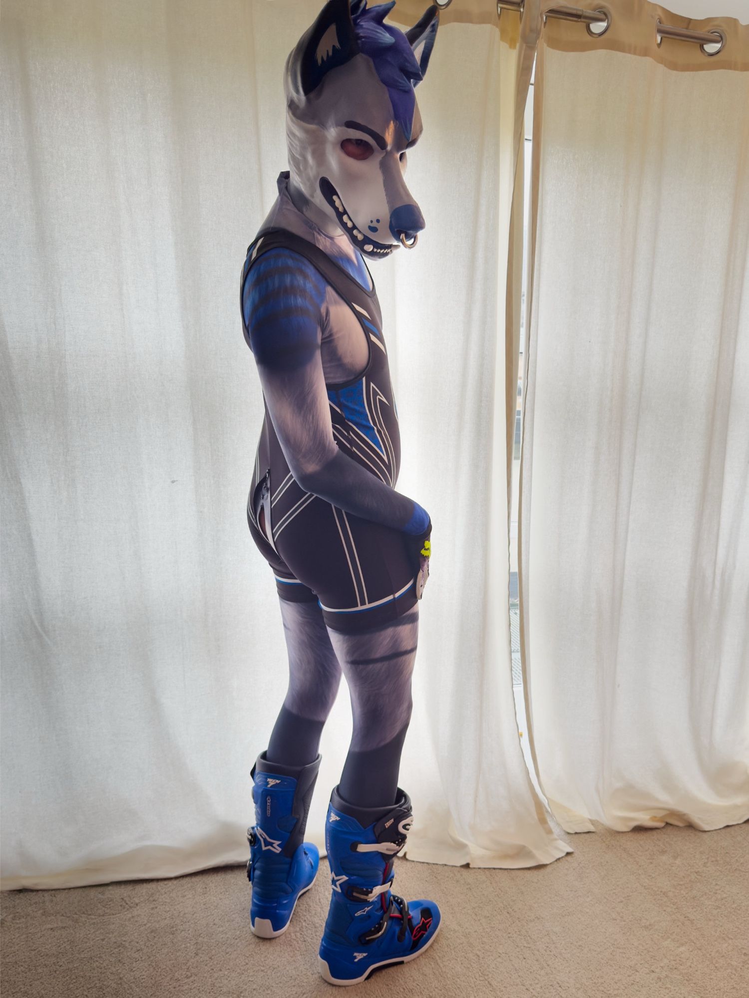 Me posing in front of a window, wearing my blue rubber wolfdog mask, skintight suit, a singlet and MX boots, and looking expectantly over my shoulder