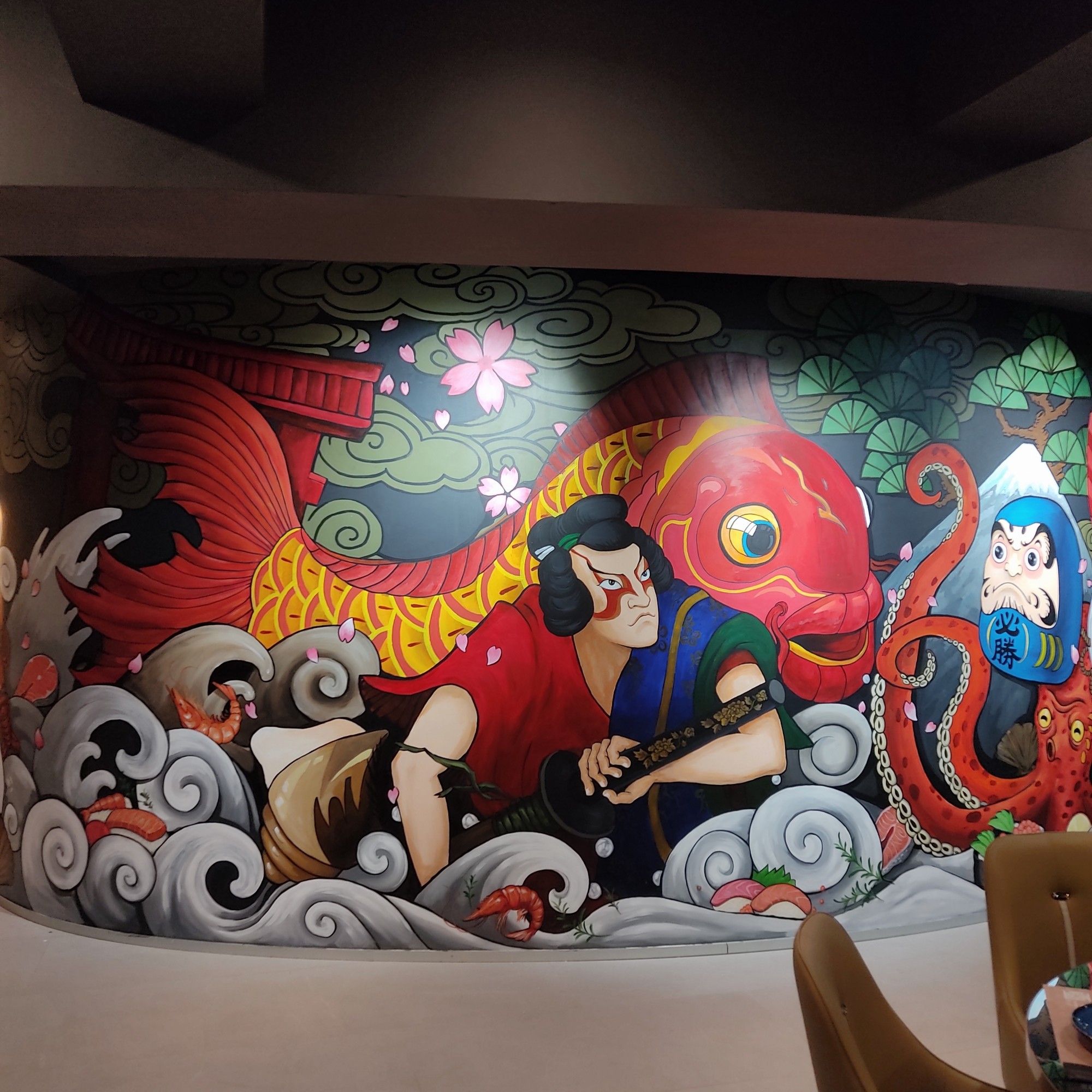 Mural de imitación japonesa en el restaurante donde se ve un samurái en el centro con una enorme carpa koi detrás. A la derecha se aprecian 2 tentáculos de un pulpo que asoma y encima un daruma de color azul con ambos ojos pintados y un monte Fuji al fondo. En la base y desde la izquierda al centro hay dibujadas unas olas donde "nadan" distintos platos (sushi y gambas cocidas) y como fondo hay un estanque con nenúfares.